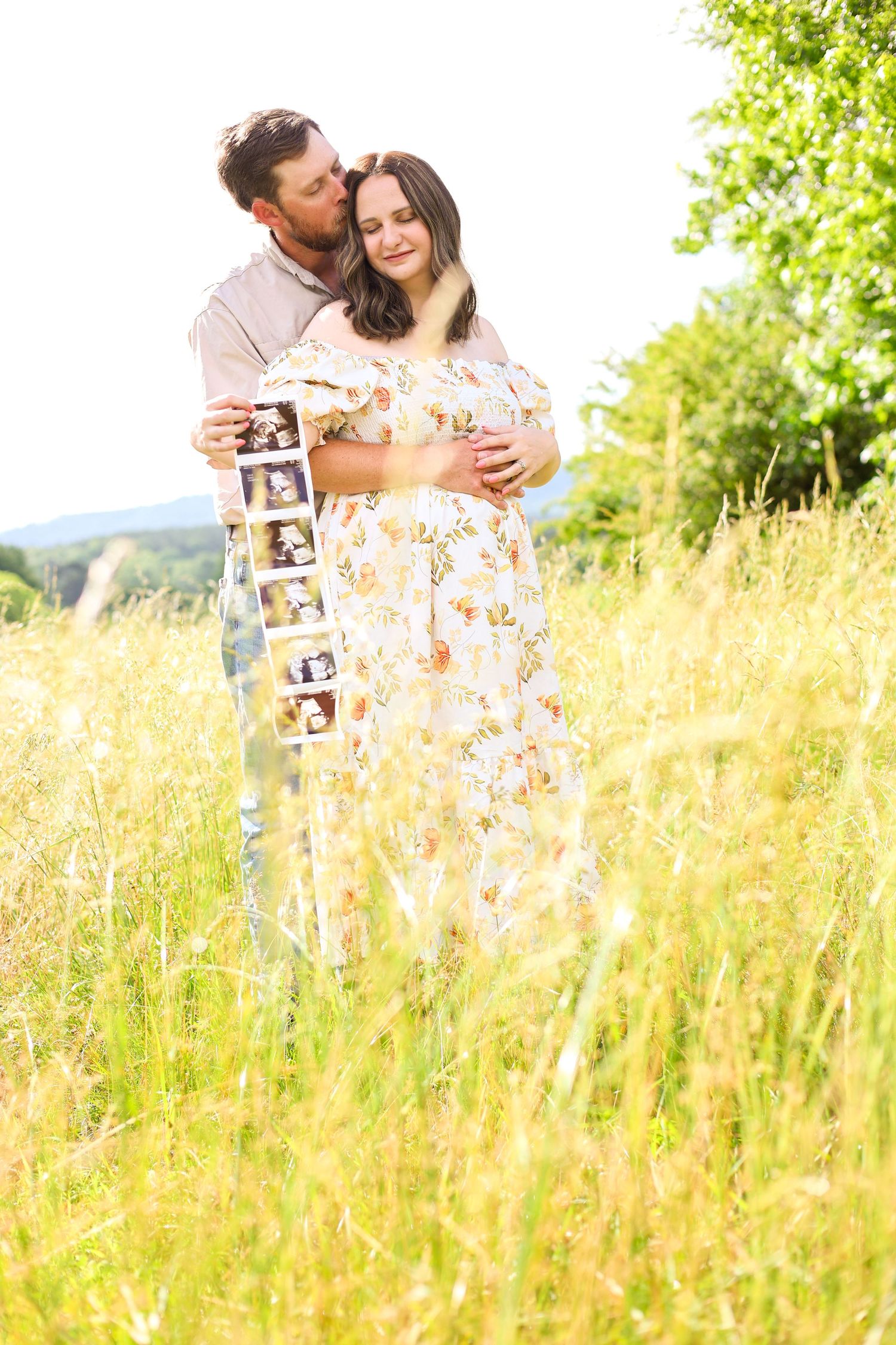 Couple embracing in a sunlit meadow of tall golden grass holding ultrasound photos.
