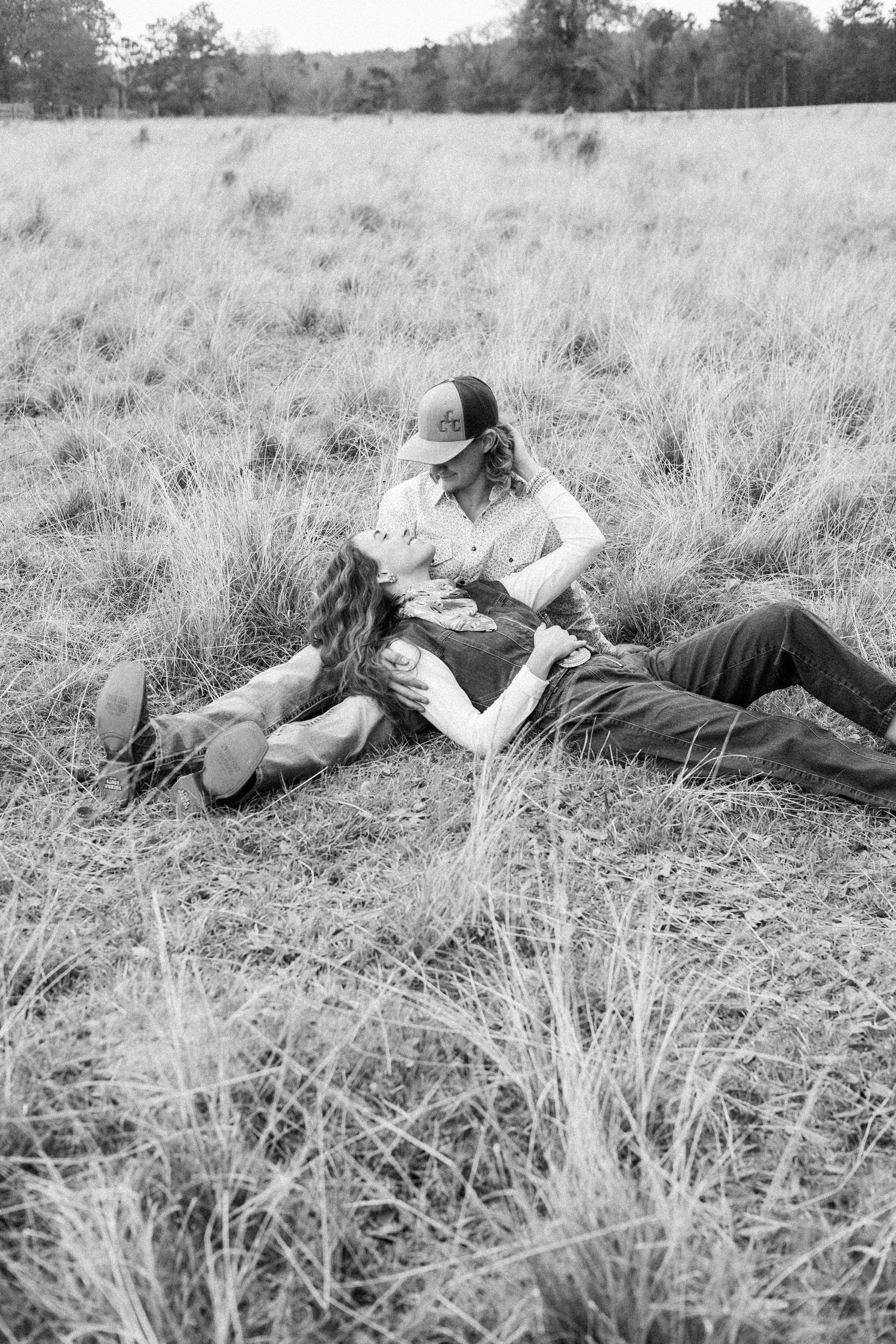 Black and white photo of a romantic couple lying in a grassy field, gazing at each other tenderly.