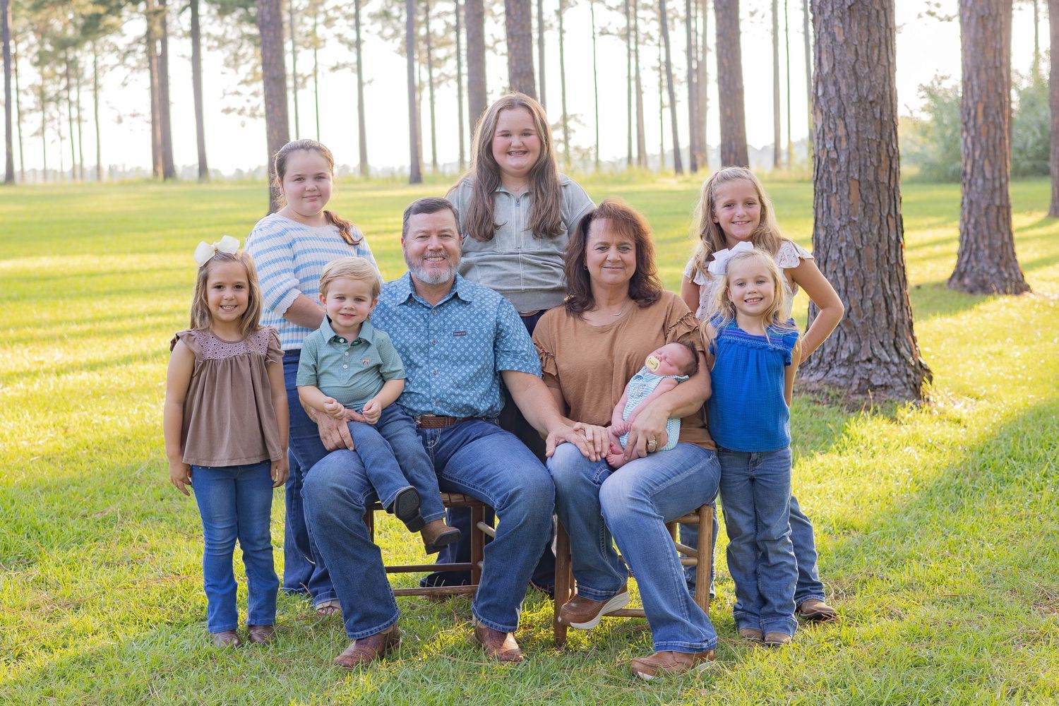Large family portrait with parents and six children posing outdoors among tall pine trees on green grass.
