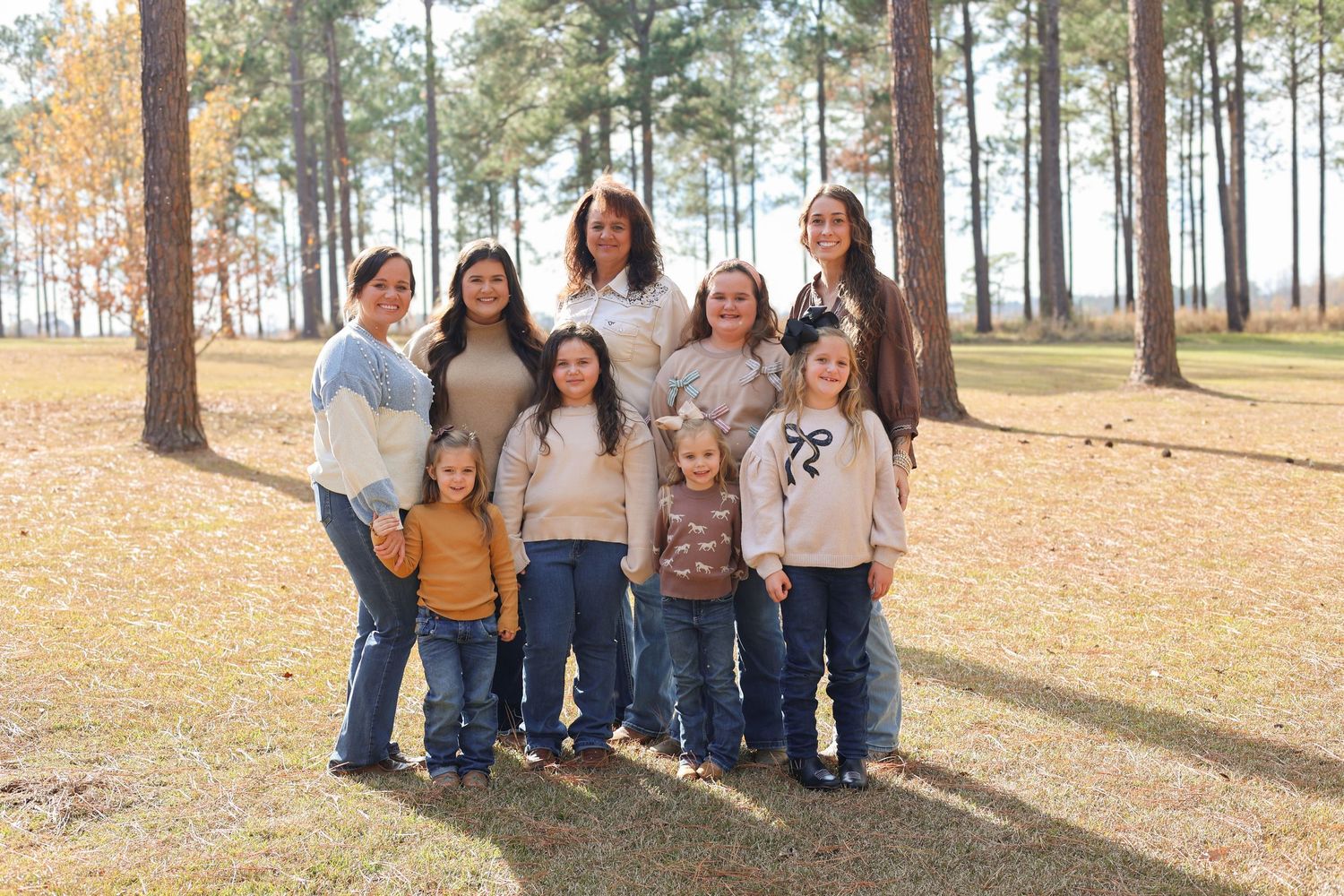 A group stands together in a casual outdoor setting with tall pine trees and fallen leaves in the background.