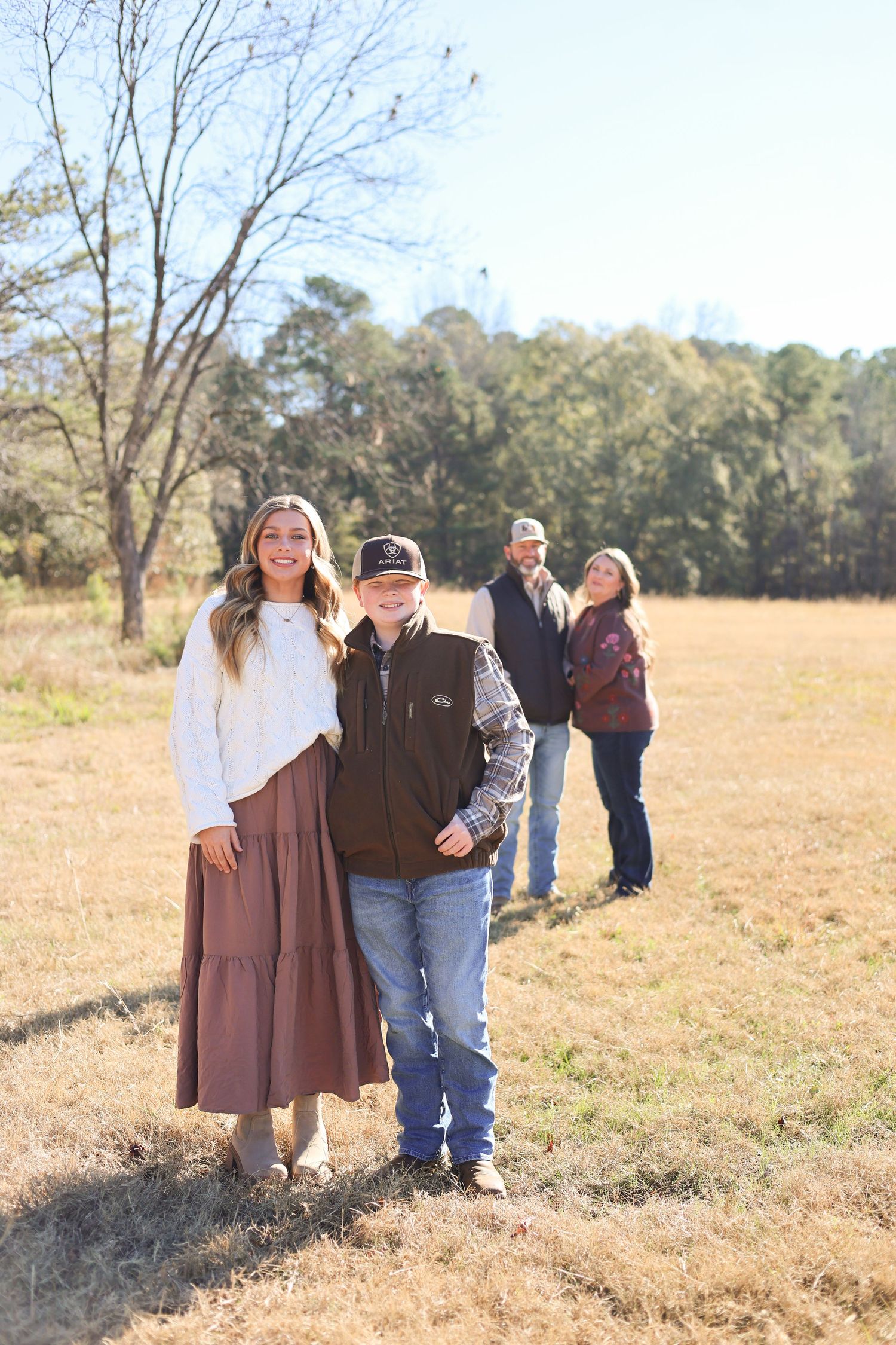 Family members standing in line in a sunny field with bare trees and natural landscape in the background.