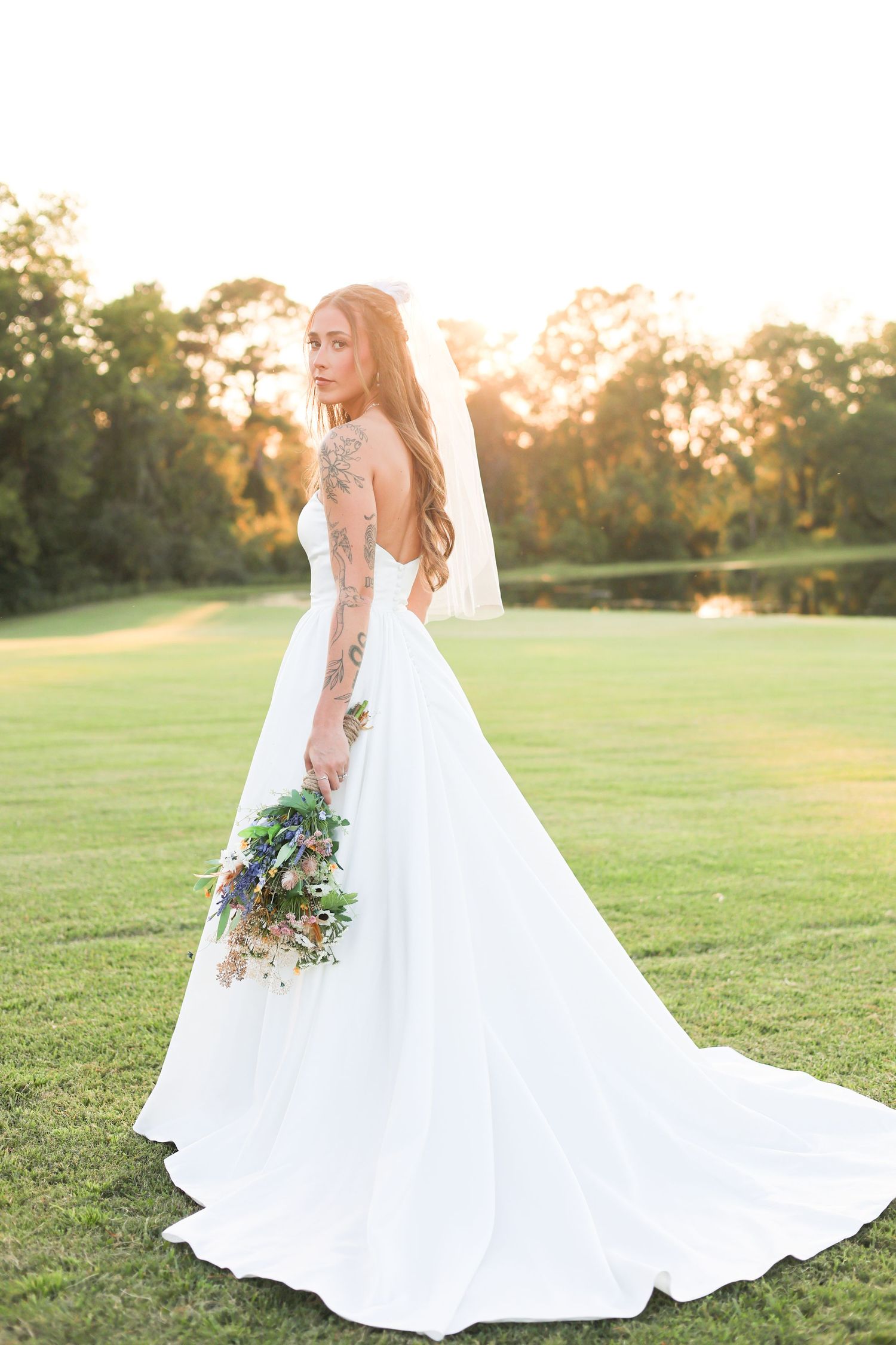 A bride in a flowing white gown with long train holds a bouquet while standing on a sunlit golf course at golden hour.