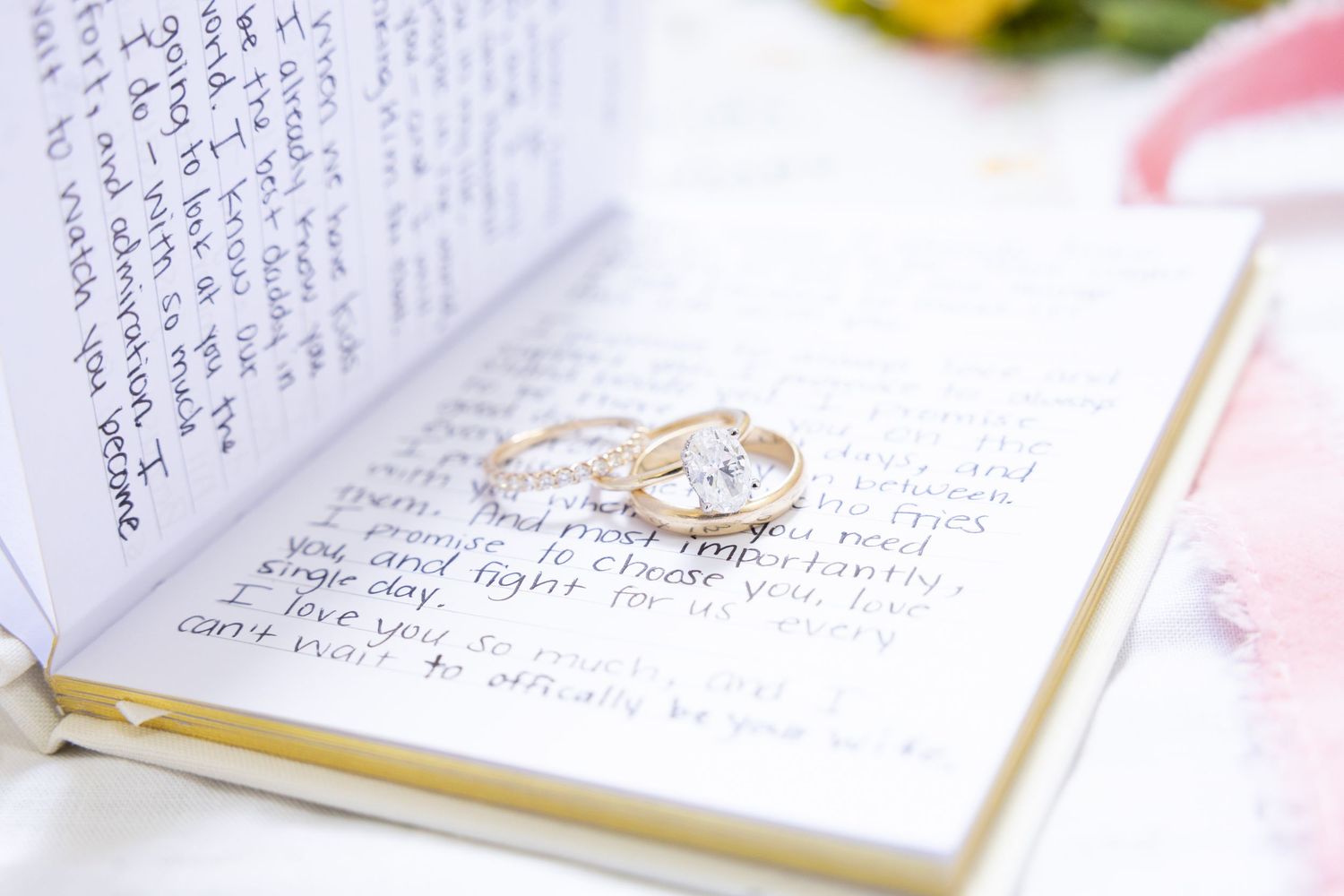 Gold wedding rings resting on an open journal with handwritten vows, with flowers in the background.
