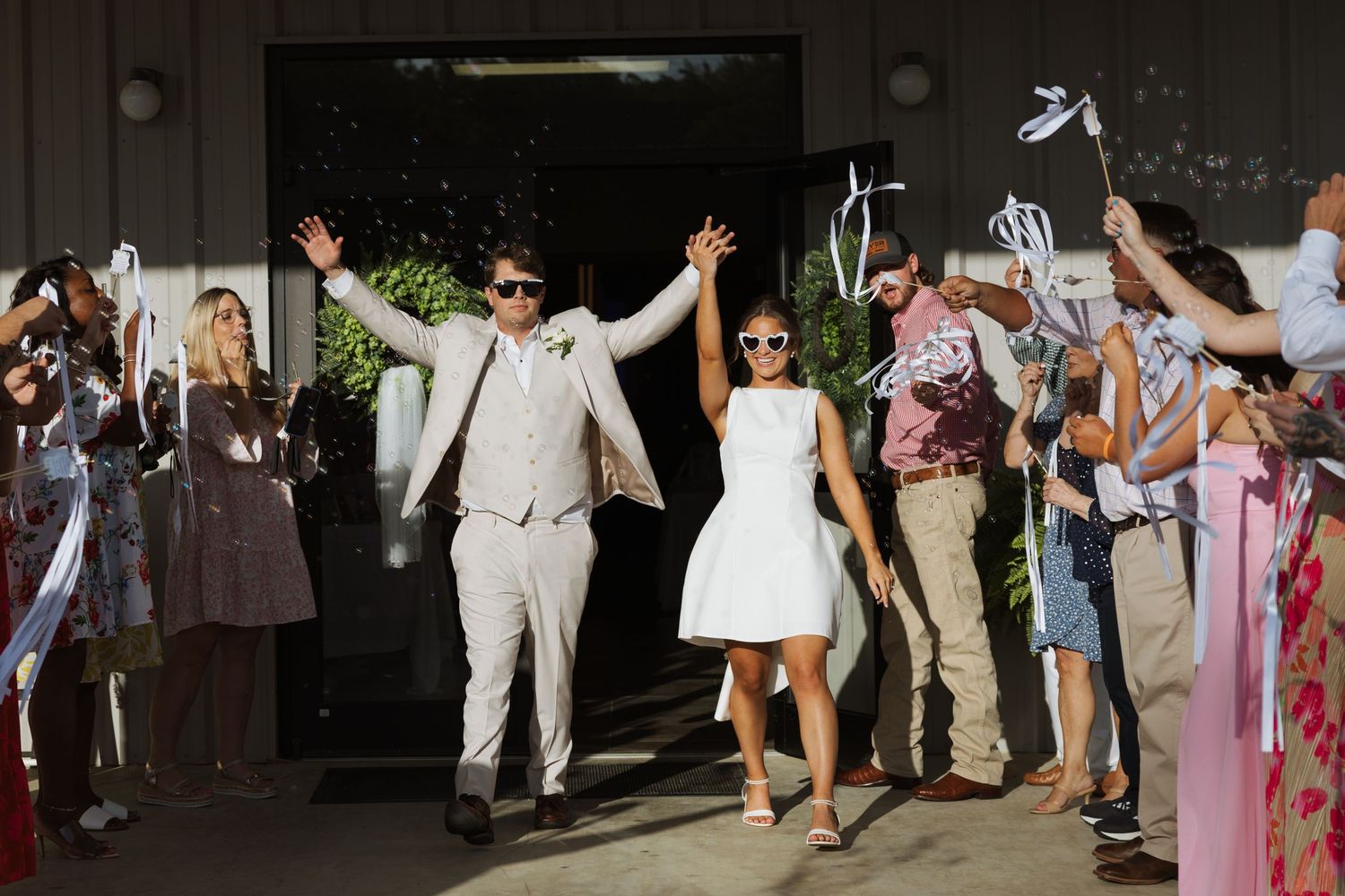 Joyful newlyweds exit wedding venue in sunglasses as guests wave ribbon wands in celebration.