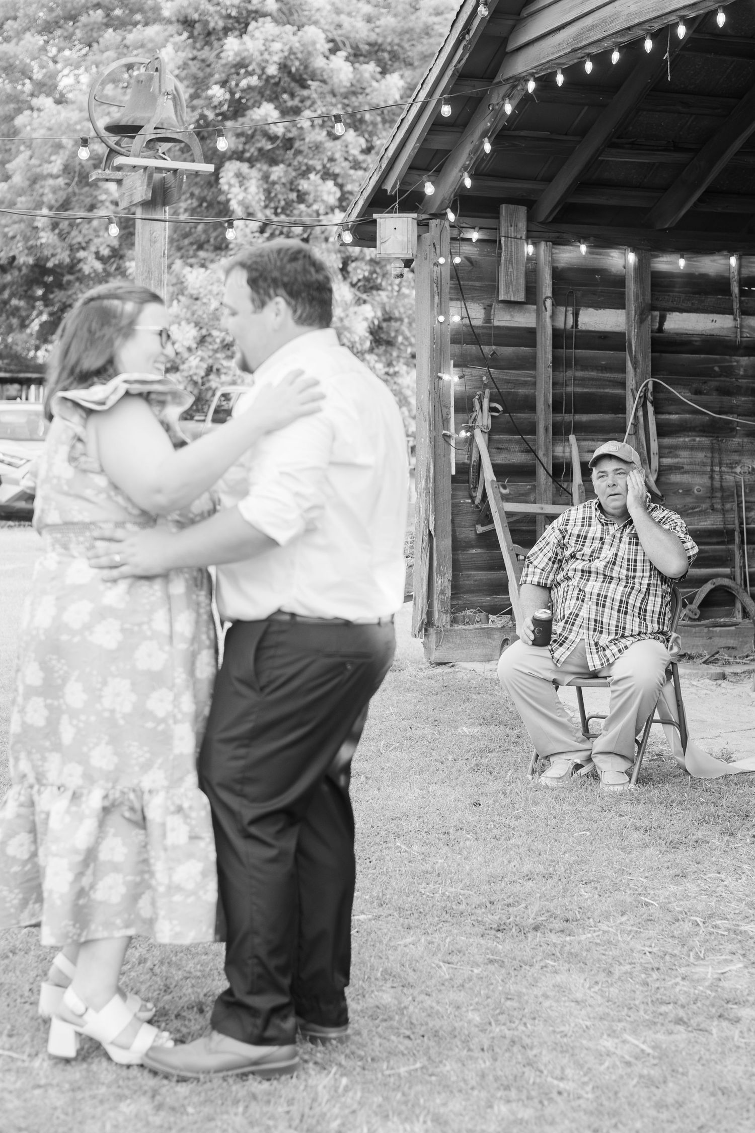 Black and white photo of a couple dancing outdoors near a rustic barn while a man sits watching nearby.