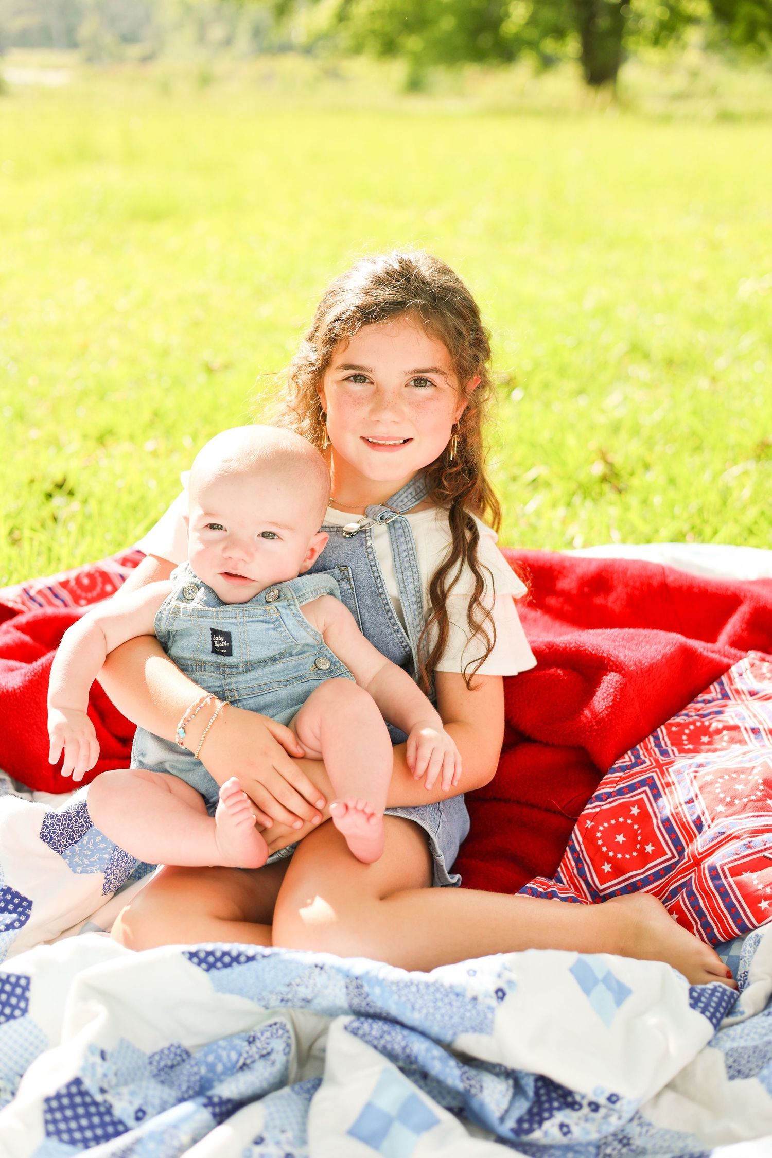 Young girl sitting on a red and blue quilt outdoors, smiling while holding a baby in denim overalls.