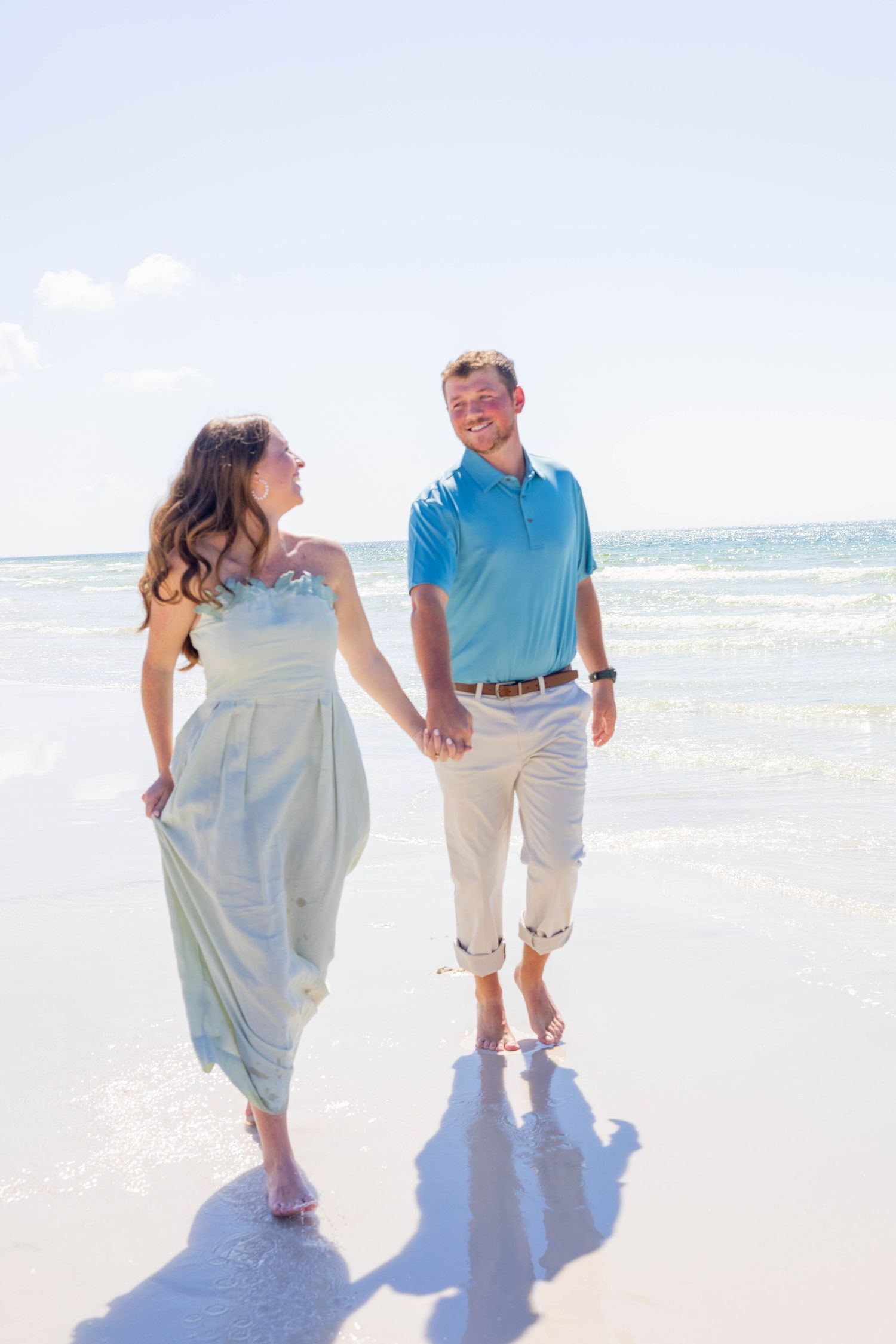 Happy couple holding hands and walking barefoot along a sunny beach, smiling at each other.