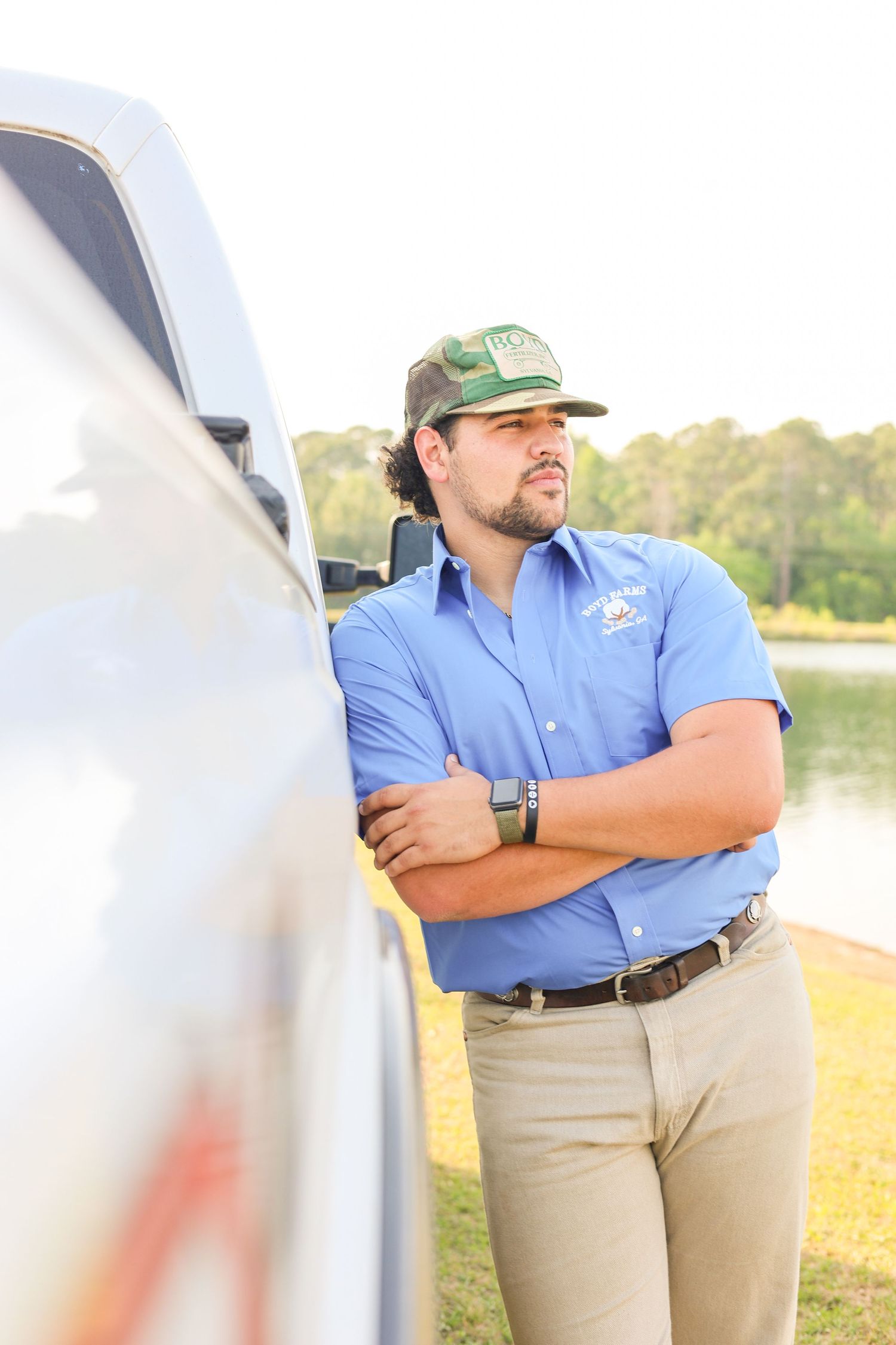 Person in blue uniform shirt and baseball cap leans against white truck near water.