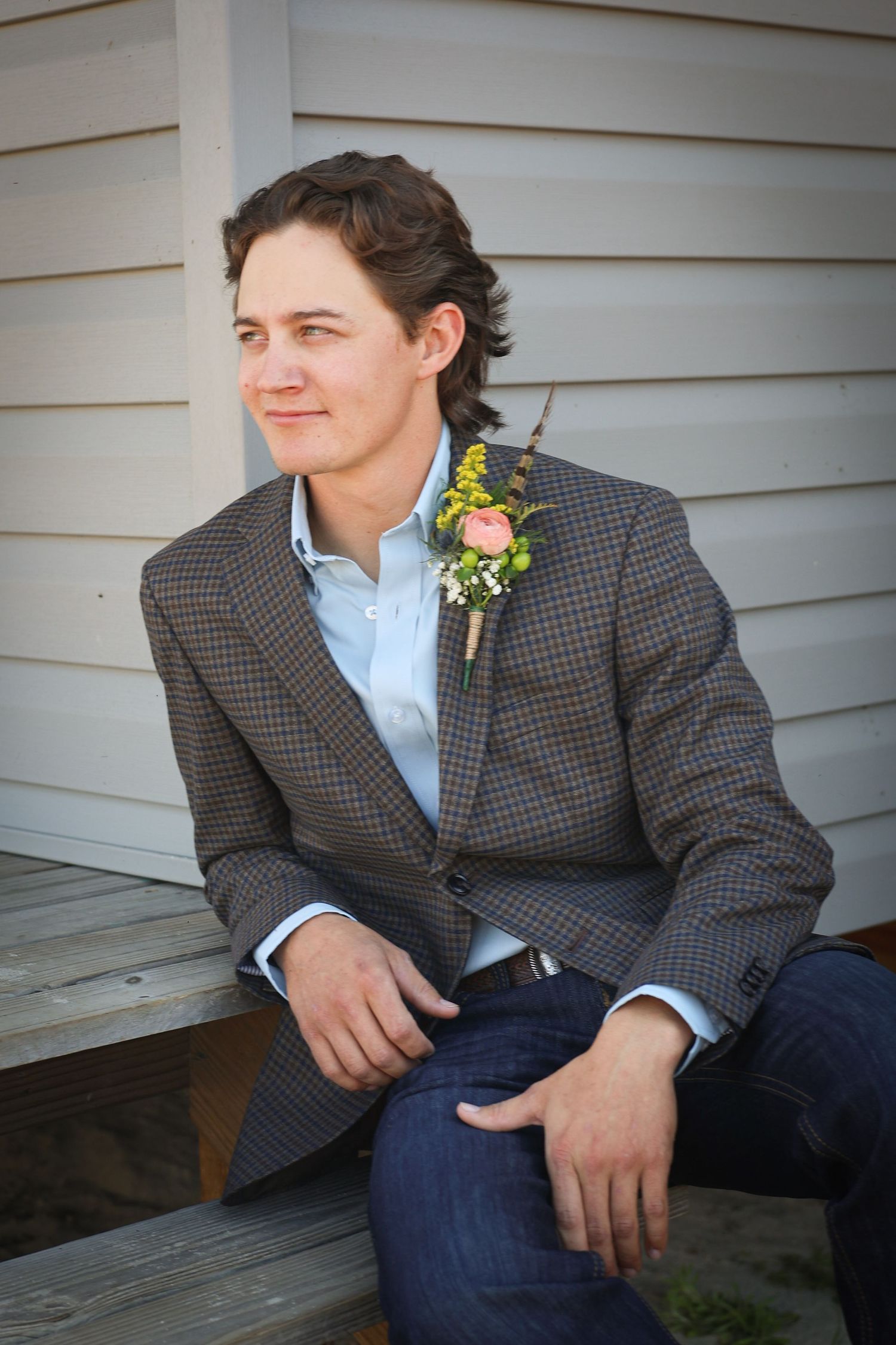 Groom in a checkered gray suit jacket and blue shirt poses on wooden steps with a floral boutonniere.