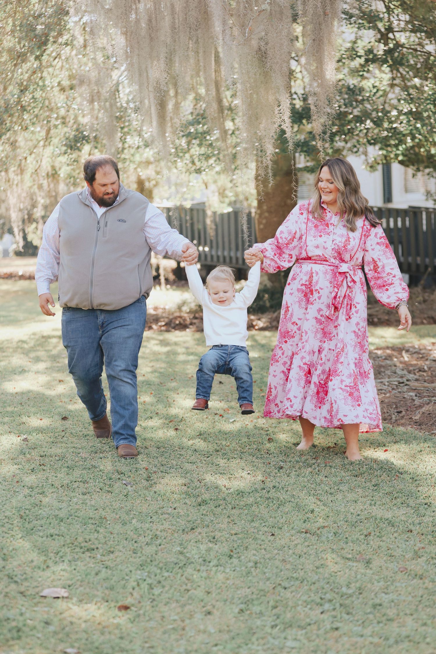 A family walks together under Spanish moss trees in a sunny garden setting.
