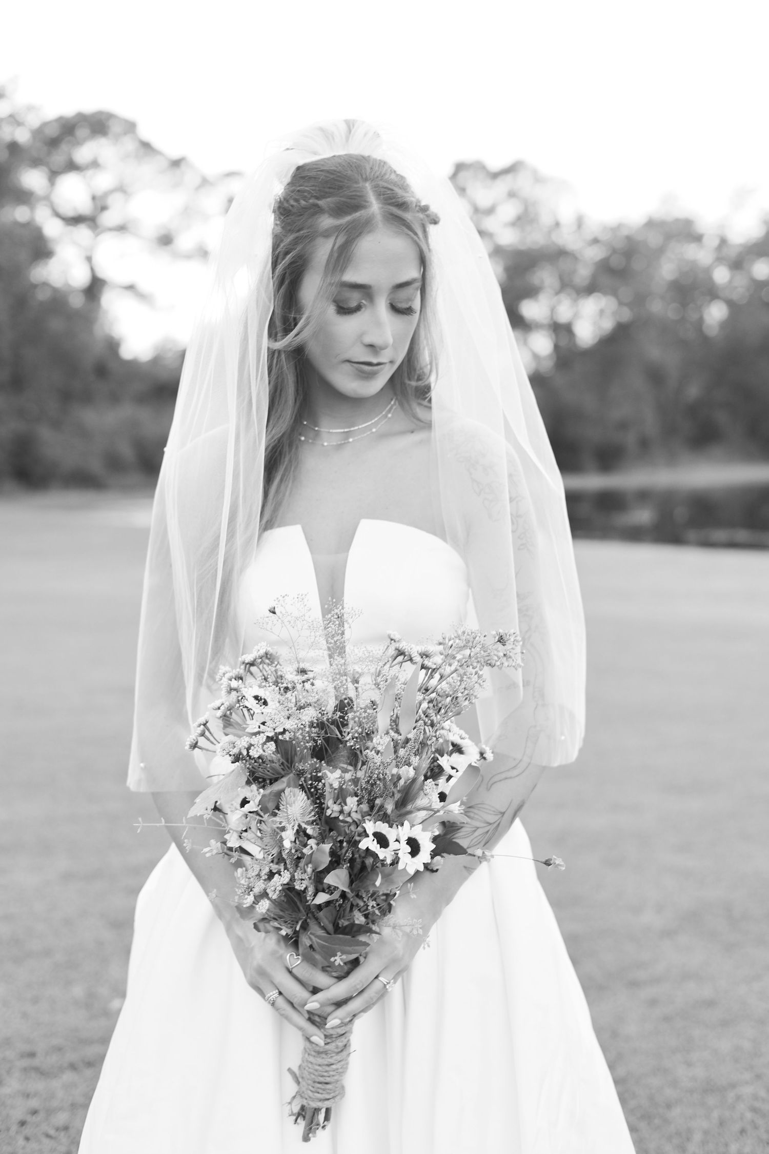 Bride in white dress holding rustic wildflower bouquet against grassy field.