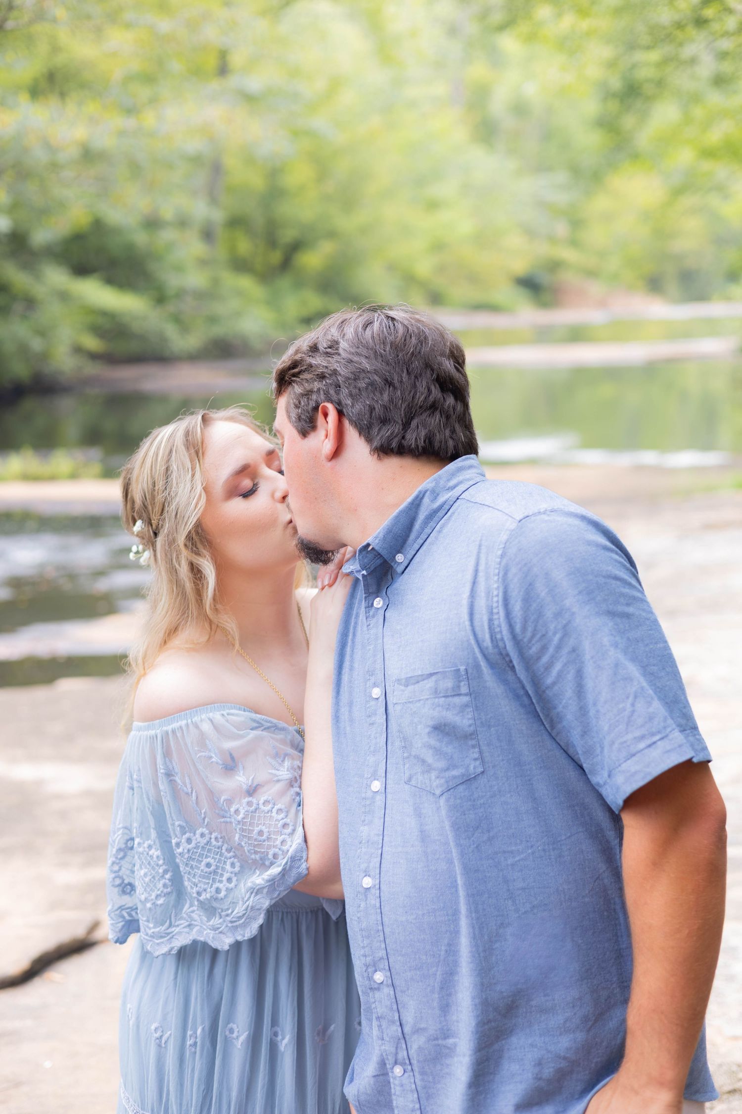 A couple shares a romantic kiss outdoors near a river, woman in blue lace dress, man in blue shirt, lush greenery behind.