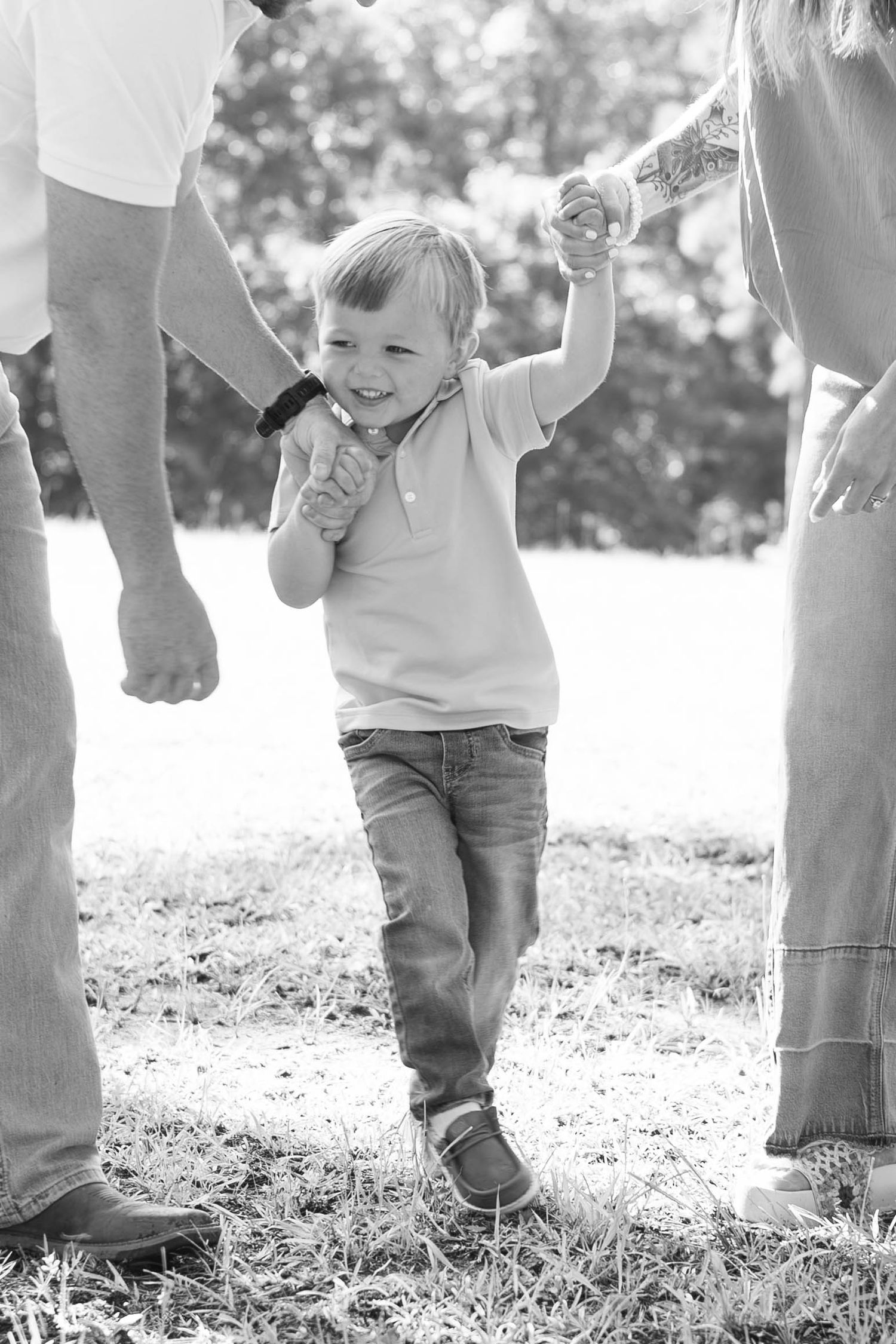 Black and white photo of a happy toddler boy holding hands with parents while walking on grass outdoors.