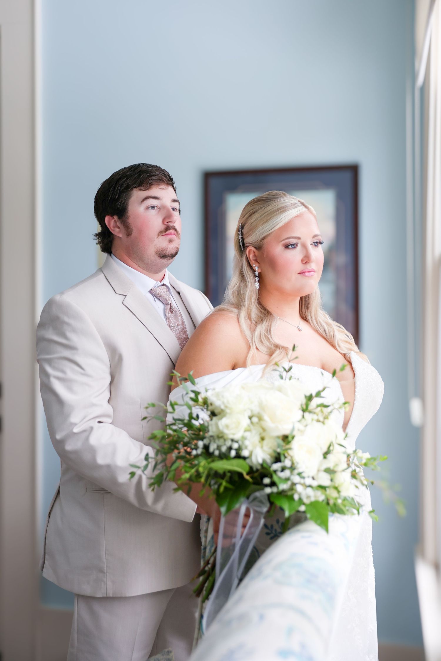 Bride and groom posing together indoors, bride holding white floral bouquet in elegant wedding attire.