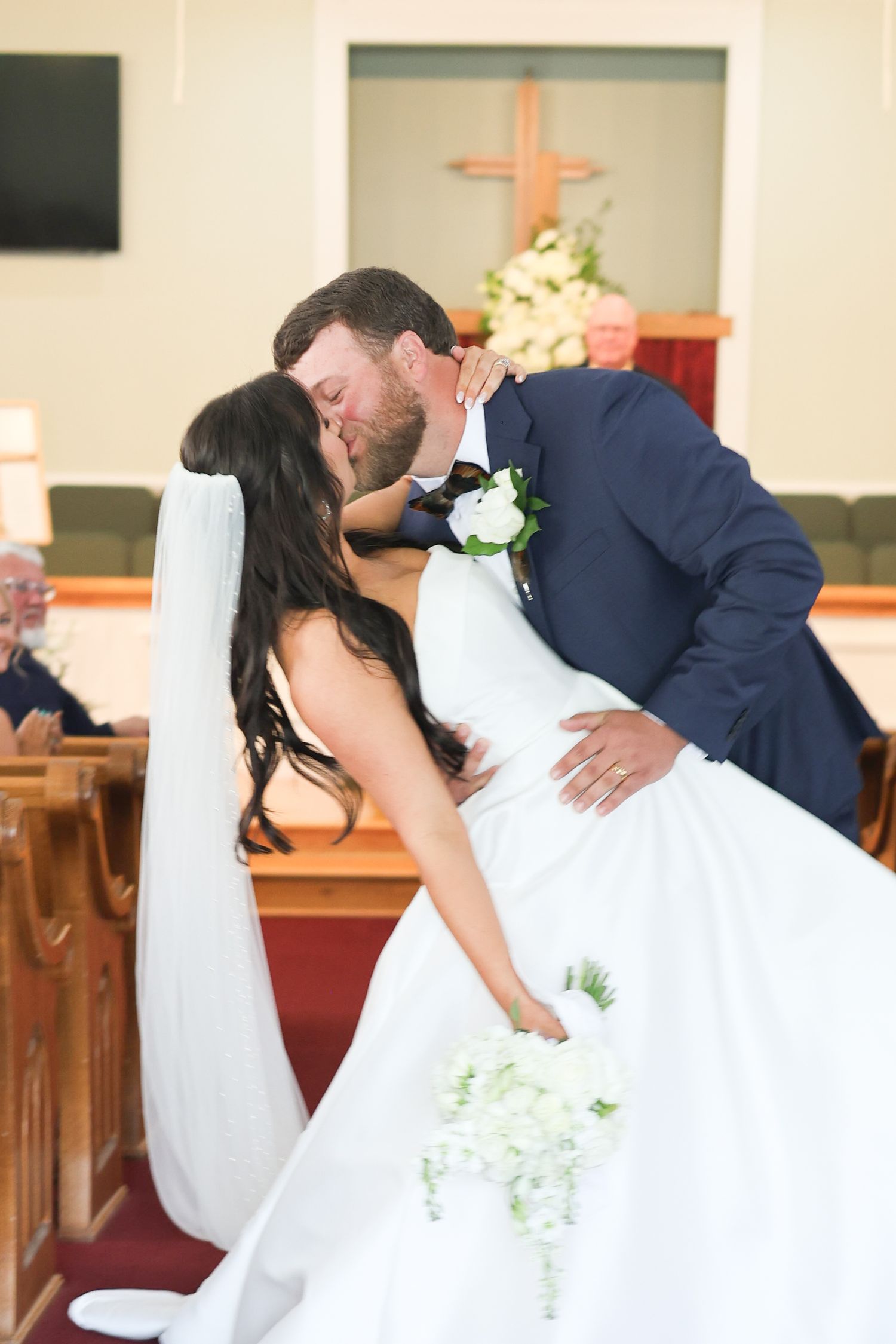 A romantic wedding kiss in a church sanctuary with a wooden cross and pews visible in the background.