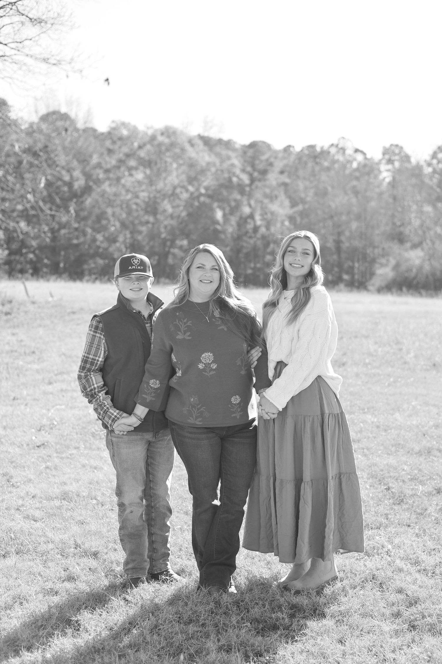 Black and white outdoor portrait of three people standing together in a grassy field with trees in the background.