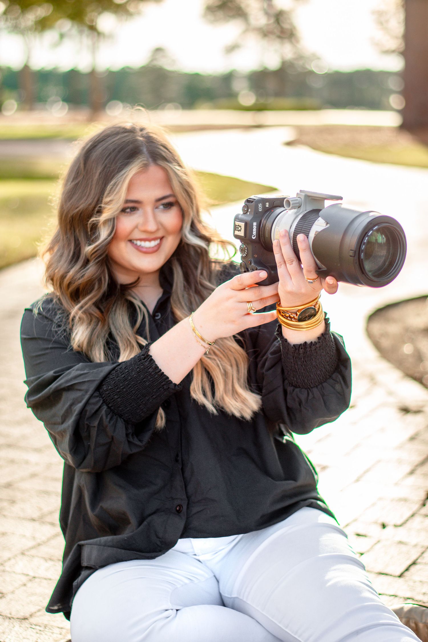 Smiling young woman with blonde highlights holding a DSLR camera outdoors, wearing a black top and white jeans.