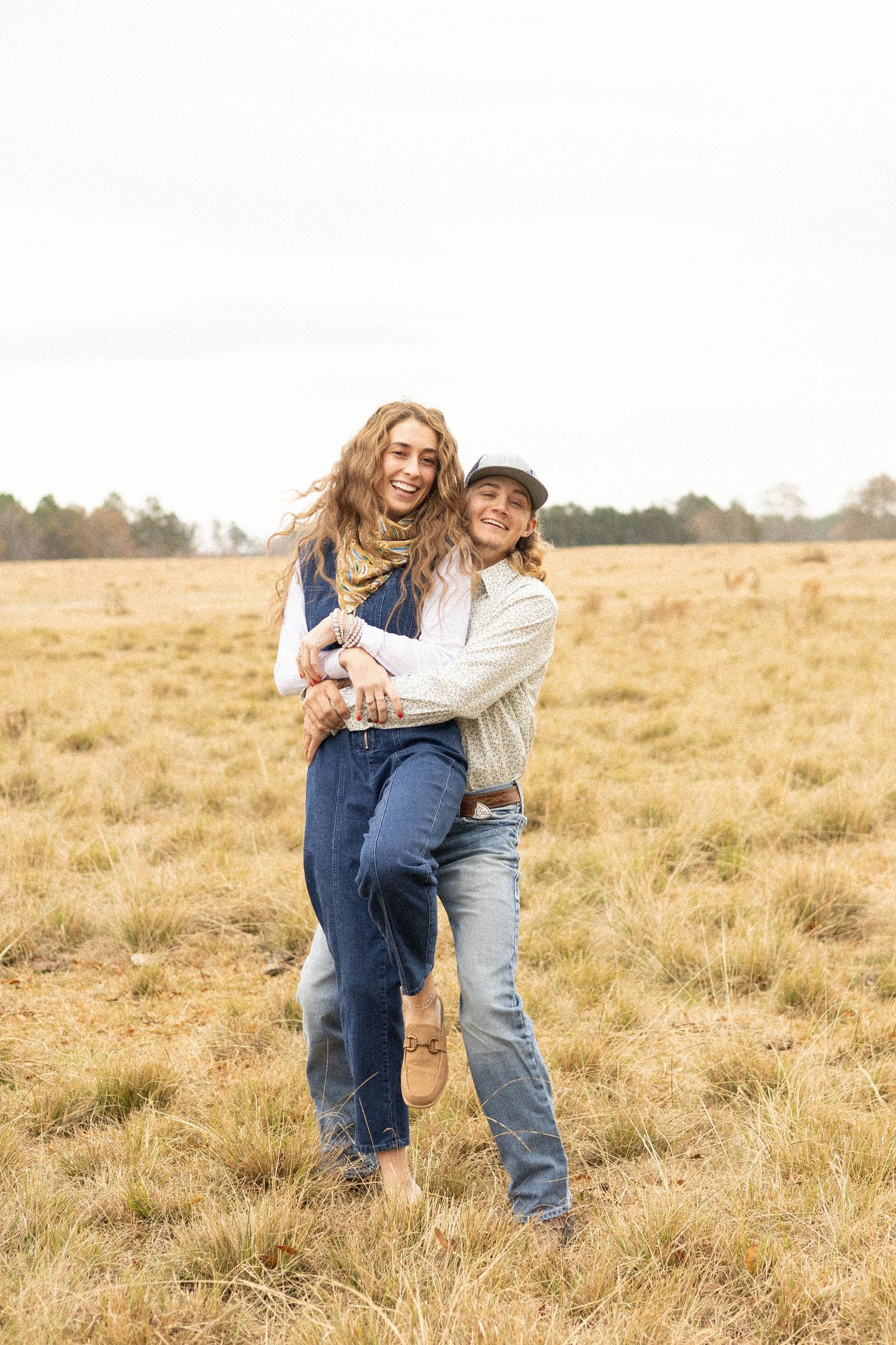 Two women embrace and laugh in a golden field, wearing denim outfits and casual layers on an overcast day.