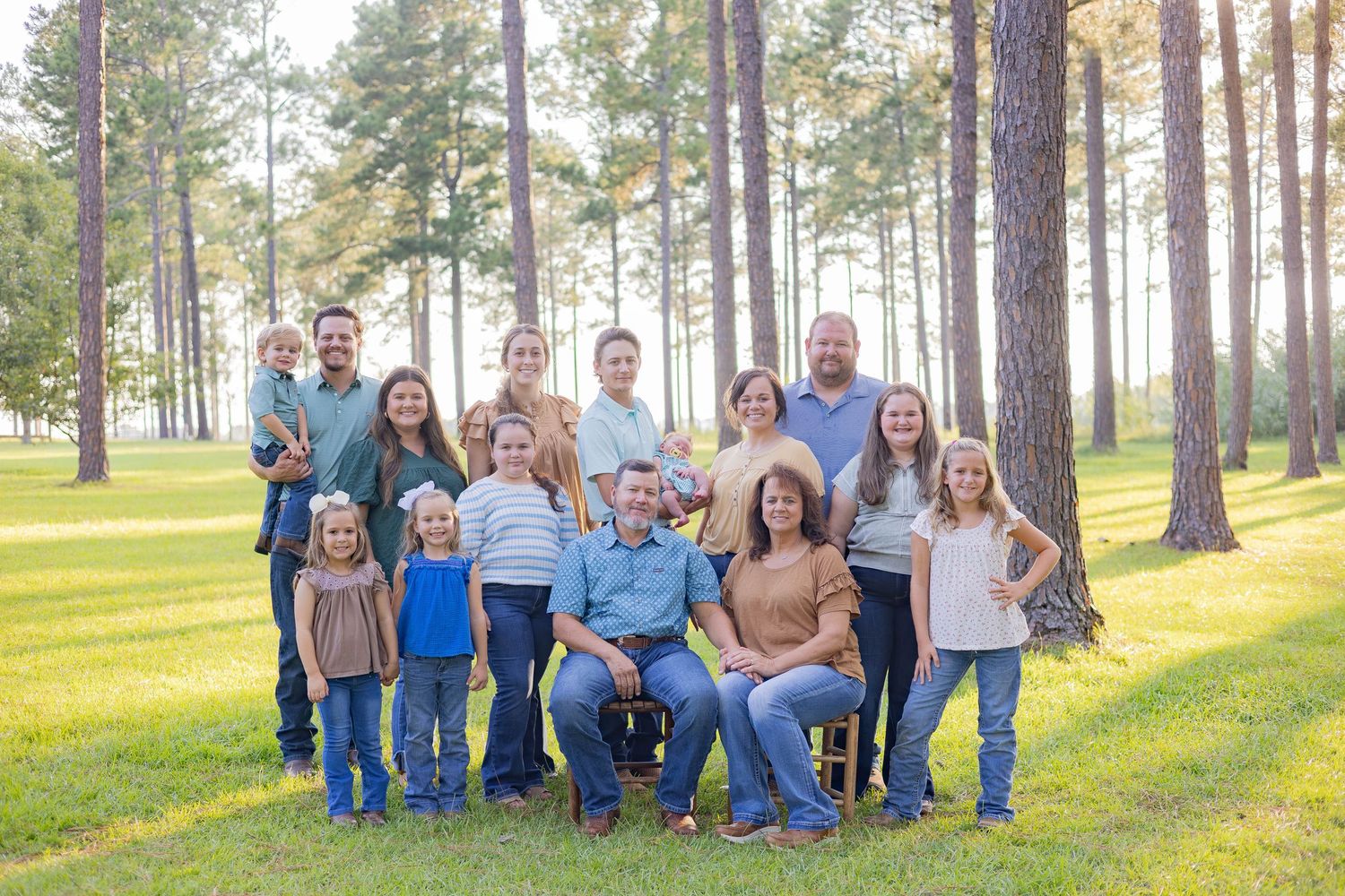 Large family portrait outdoors among tall pine trees, posing together on green grass in casual attire.