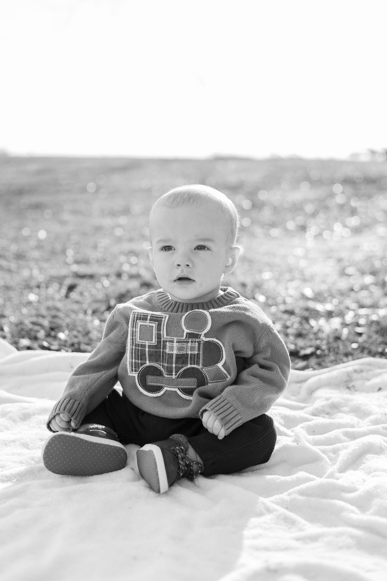 Black and white photo of a baby boy sitting on a white blanket outdoors, wearing a train sweater and small shoes.