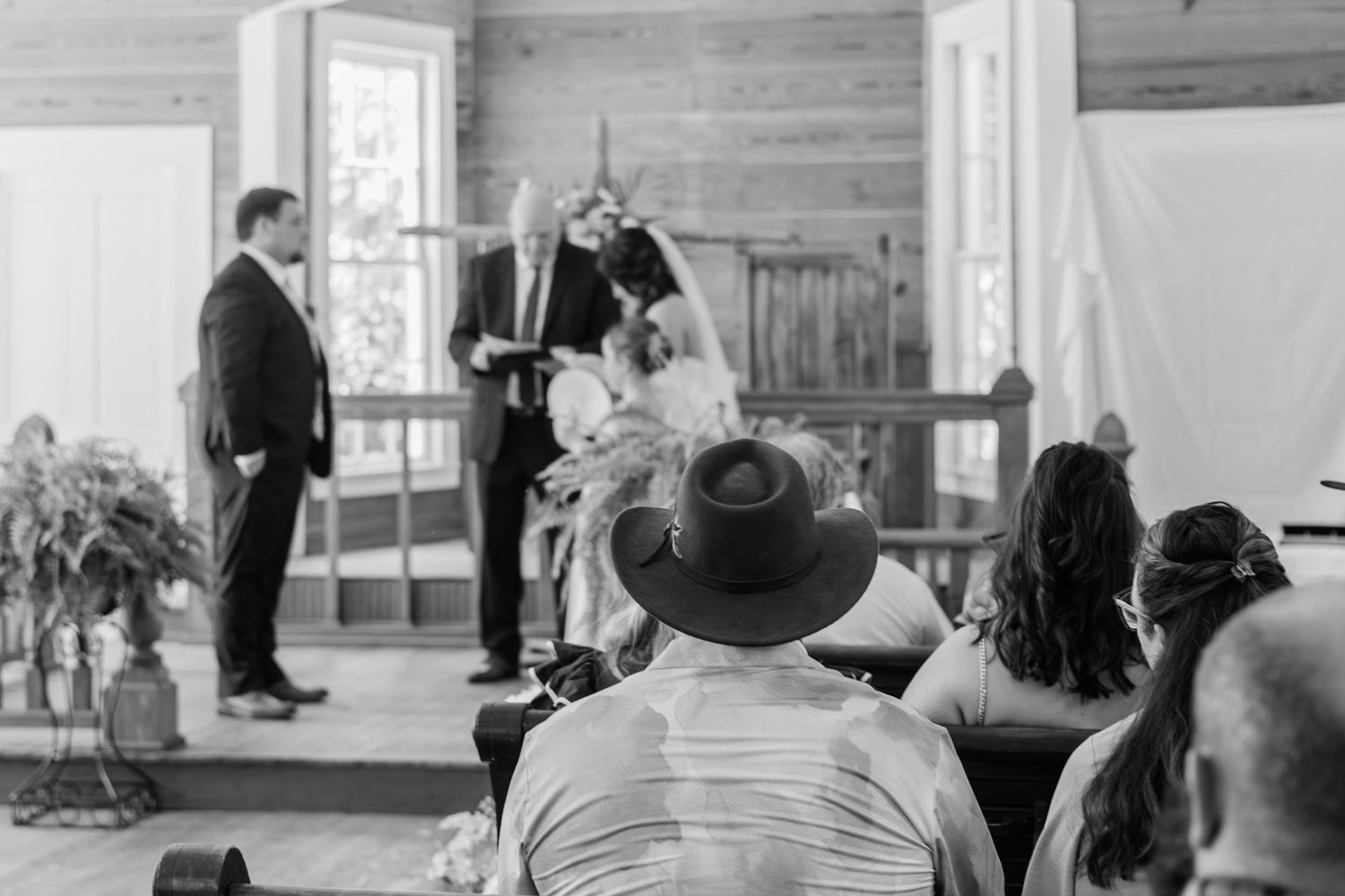 Black and white wedding ceremony photo with guests seated watching couple exchange vows at rustic wooden altar.