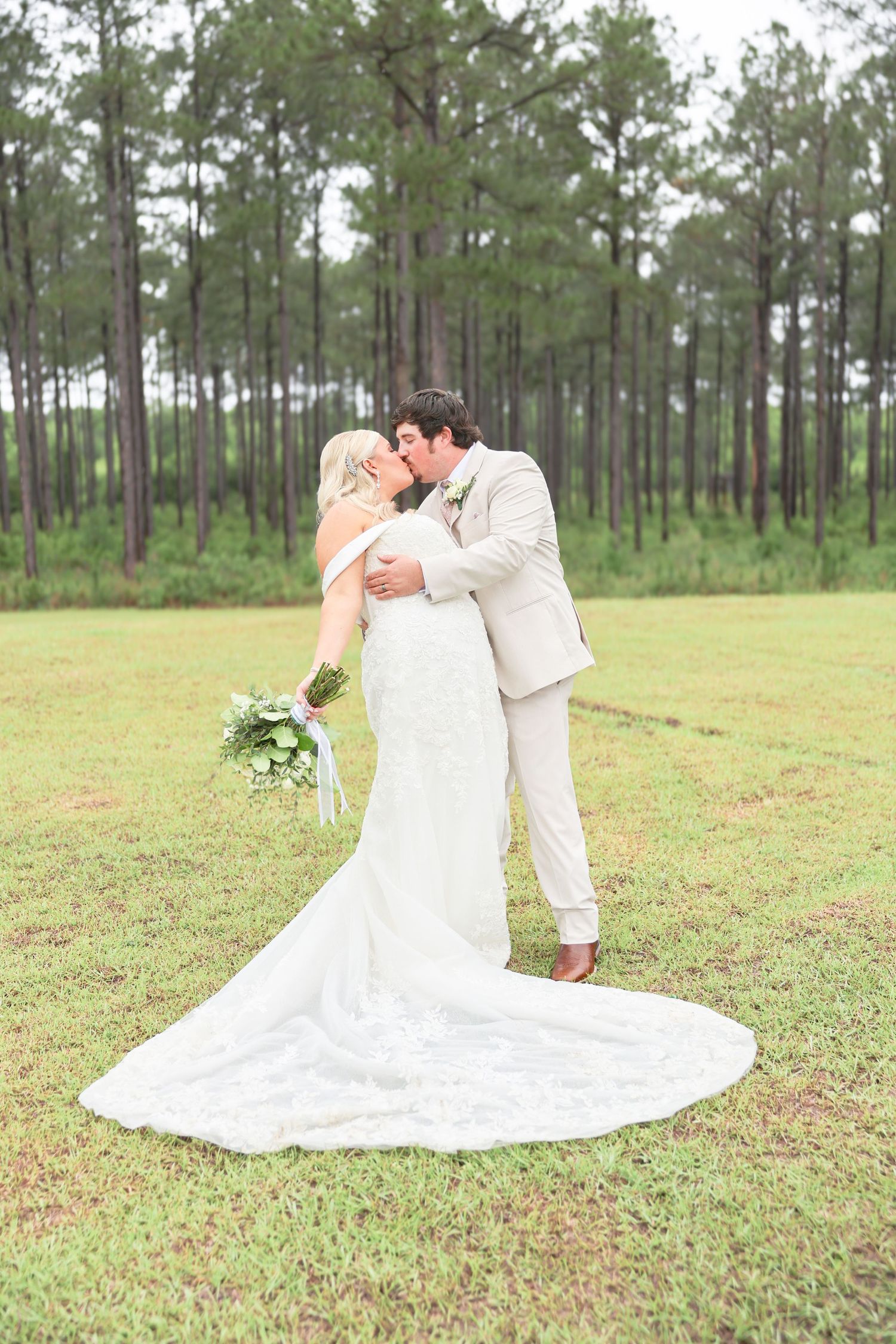 Bride and groom share a kiss outdoors in a pine forest, bride in lace gown holding greenery bouquet.