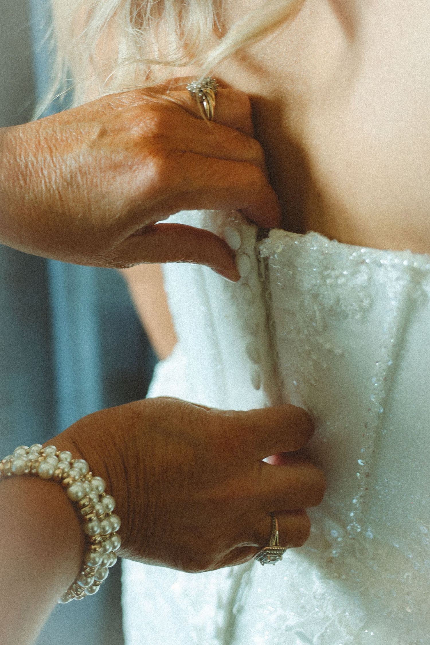 Hands fastening a white lace wedding dress on a bride, with pearl bracelet and gold rings visible.