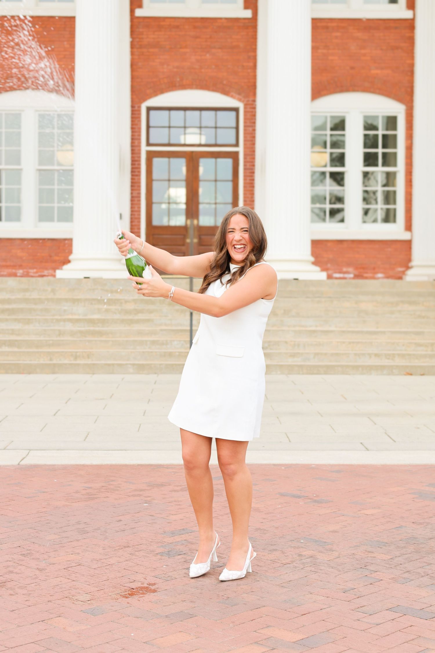 Graduate celebrates on brick courtyard in front of collegiate building.