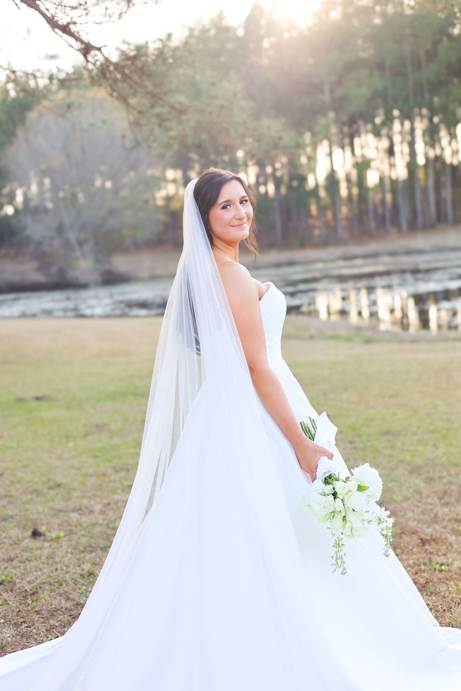 Bride in flowing white ballgown poses in golden sunlight among tall pine trees.