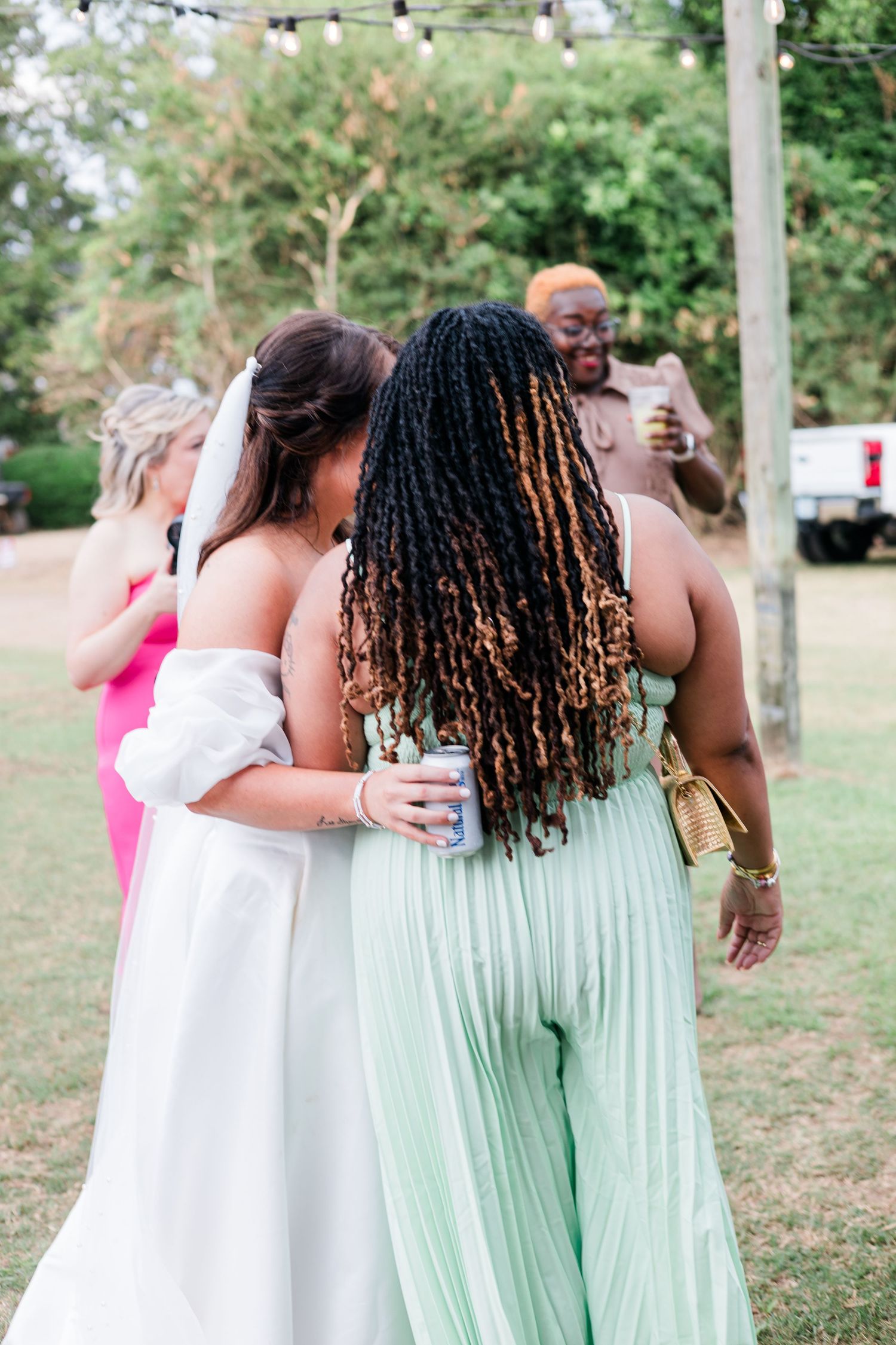 Bride in white gown embraces guests in mint green outfit at an outdoor wedding reception.