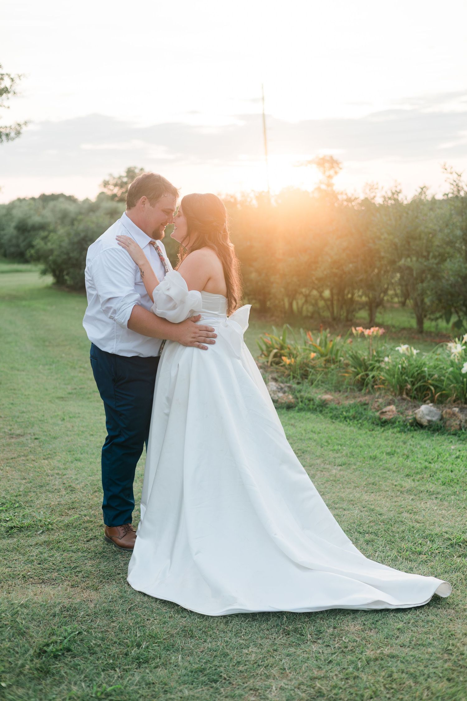 Bride and groom embracing at golden hour sunset in a lush green garden, bride wearing elegant white gown with train.