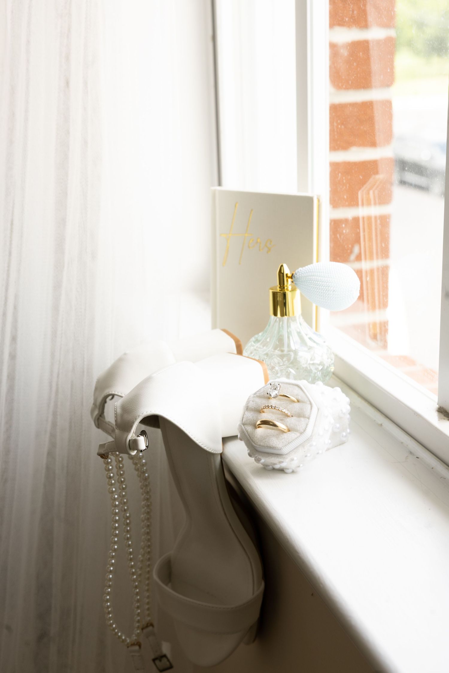 Bridal accessories including white heels, pearl necklace, and perfume on windowsill.