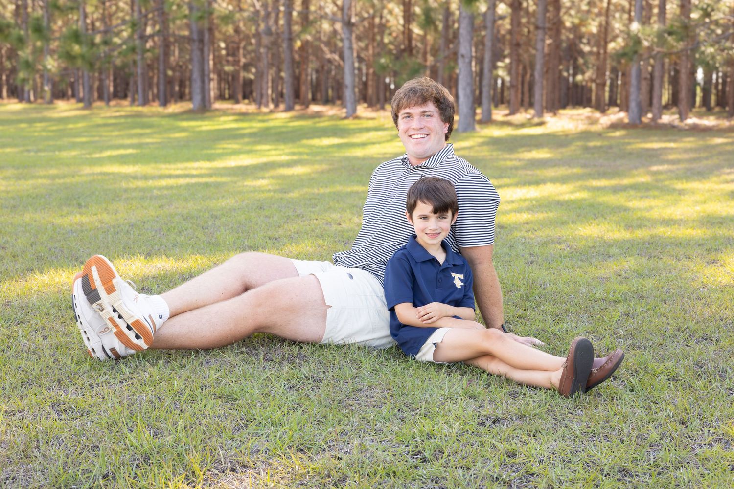Two boys smiling and relaxing together on green grass with pine trees in the background during golden hour.