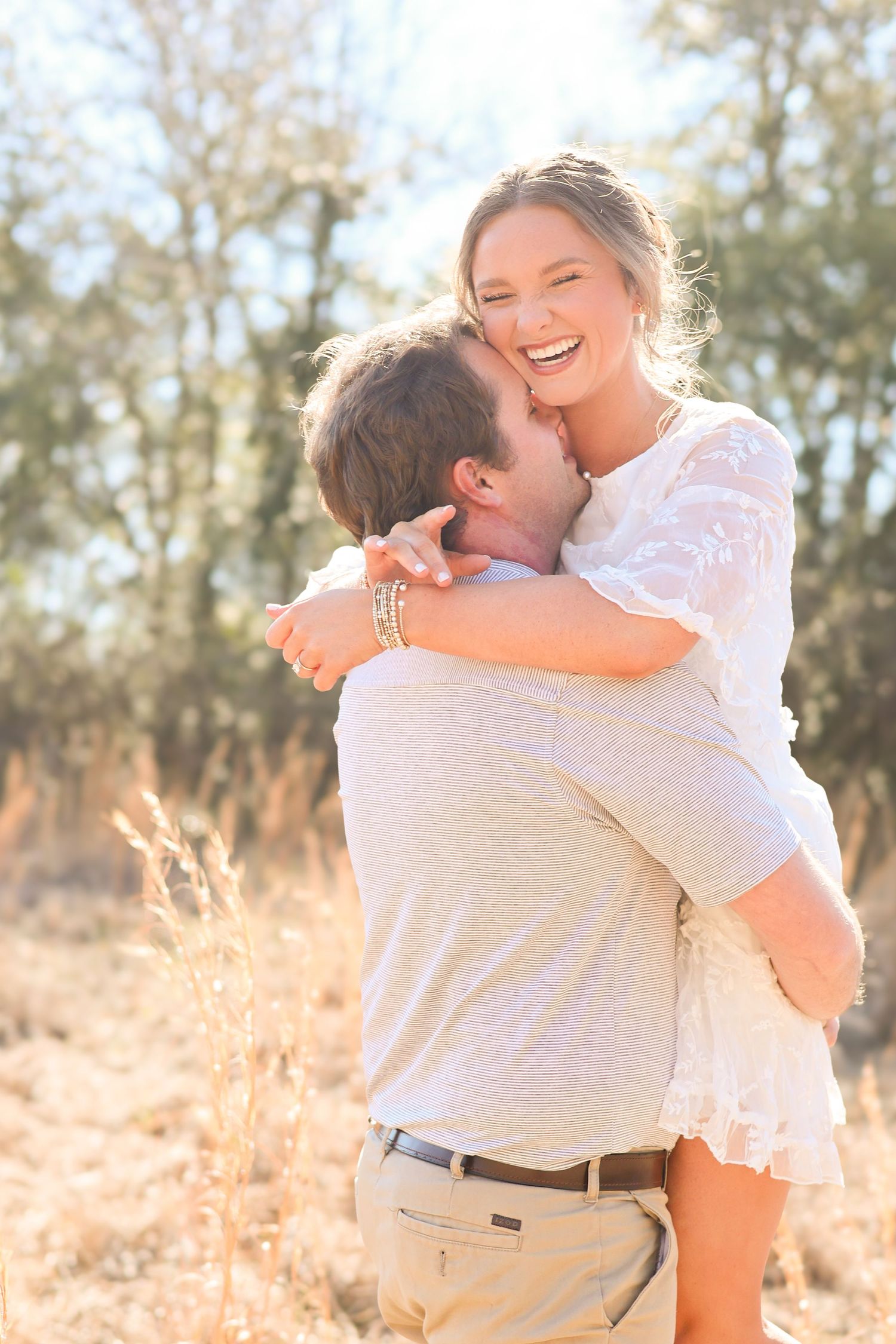 A couple embraces joyfully in a sun-drenched field with golden grass and trees in the background.