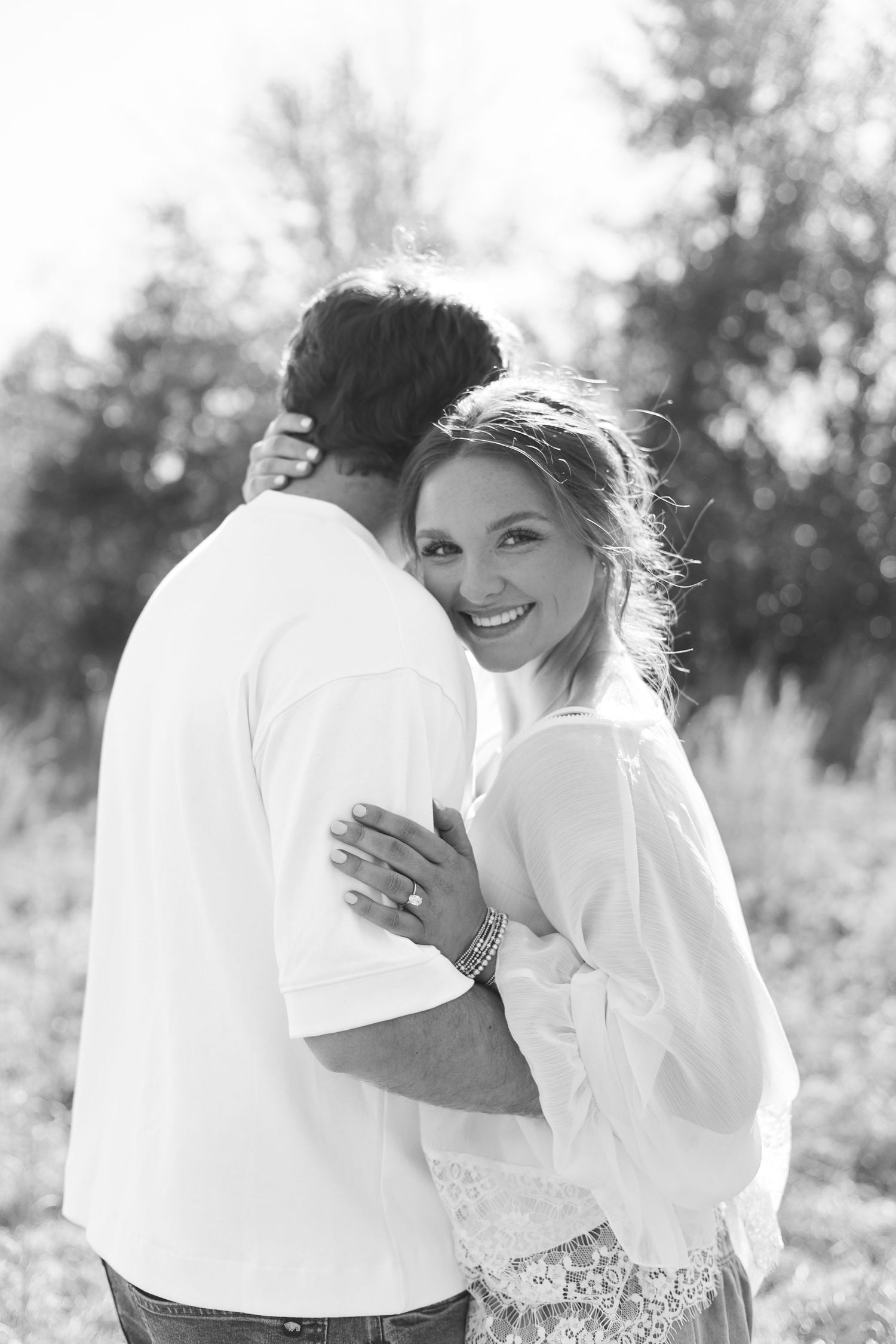 Black and white romantic photo of a couple embracing outdoors with sunlit trees in the background.