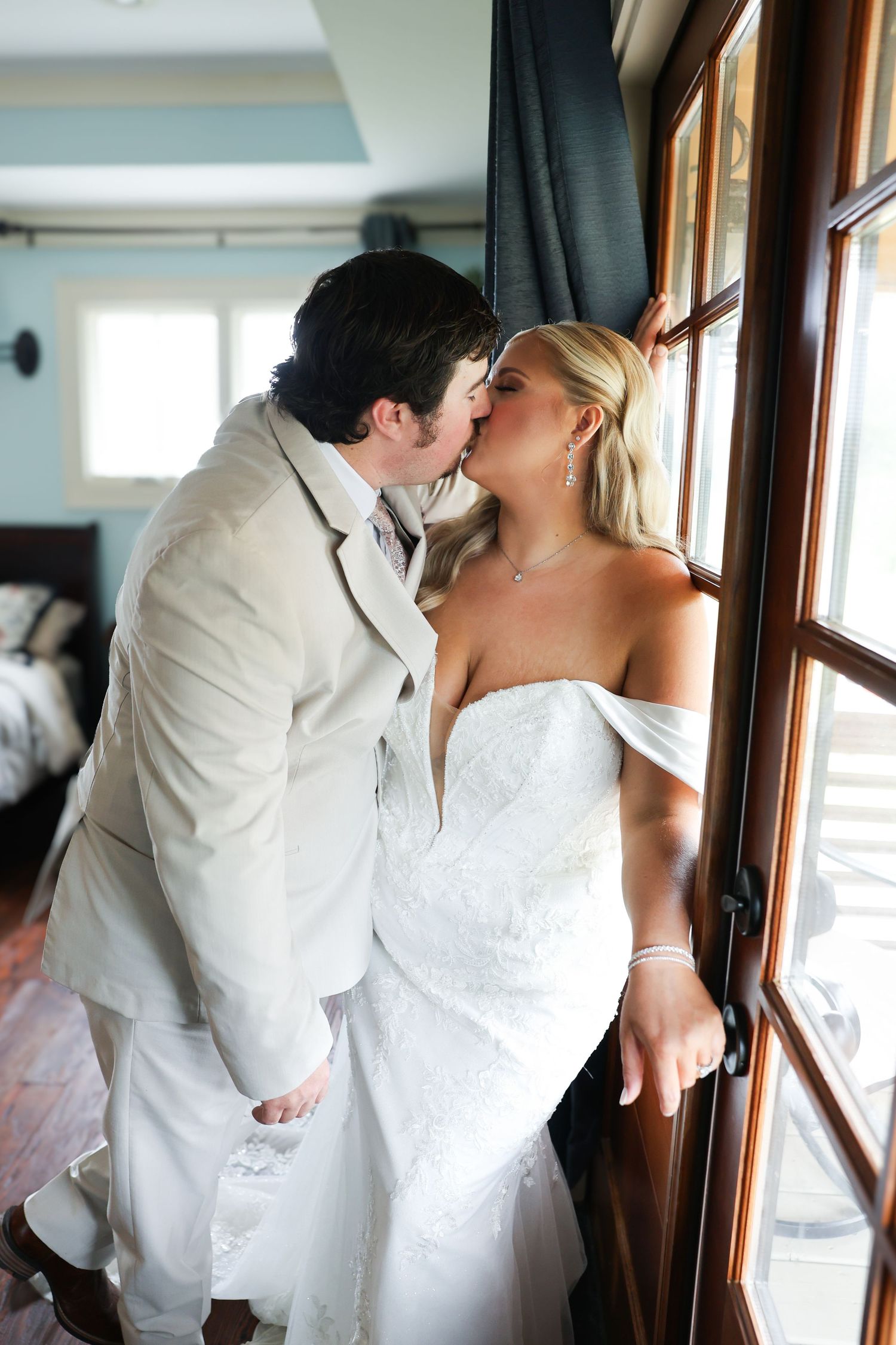 A bride and groom share a romantic kiss by a sunlit window, dressed in a white gown and beige suit on their wedding day.
