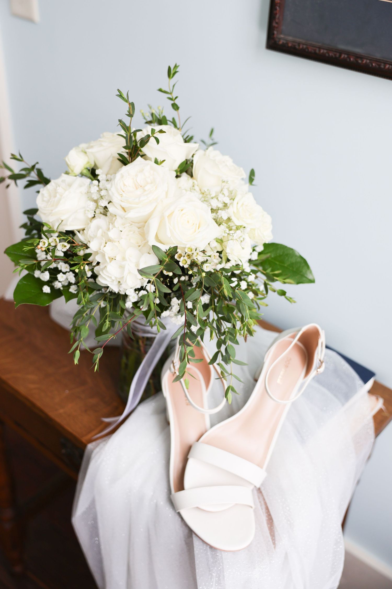 Elegant bridal flat lay featuring white rose bouquet with greenery and nude strappy heels on wooden dresser.