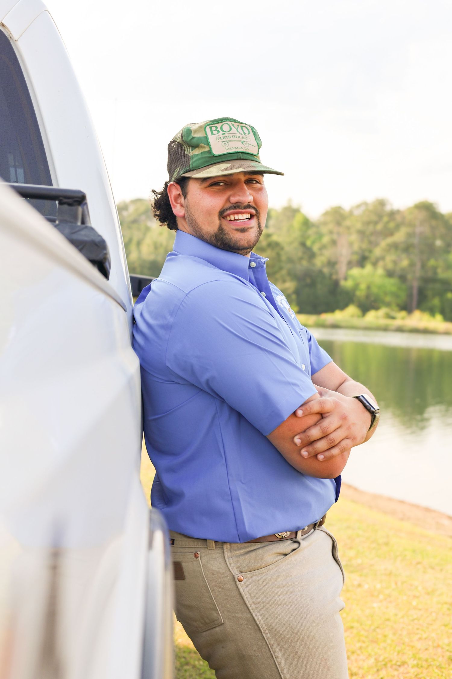 Person in blue work shirt and green cap poses beside white truck near lake.