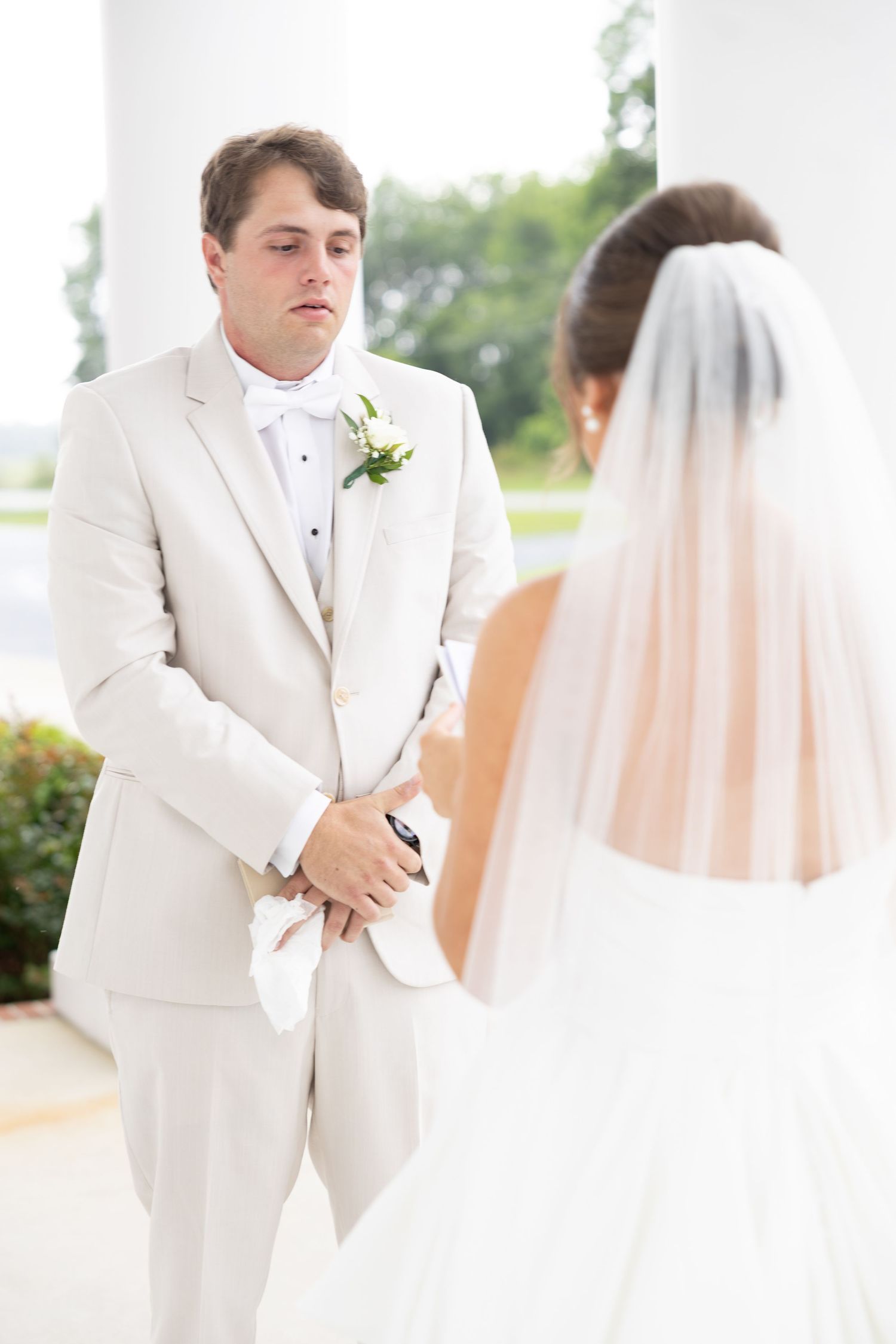 Groom in cream suit reads vows while facing bride in white dress and veil during first look.