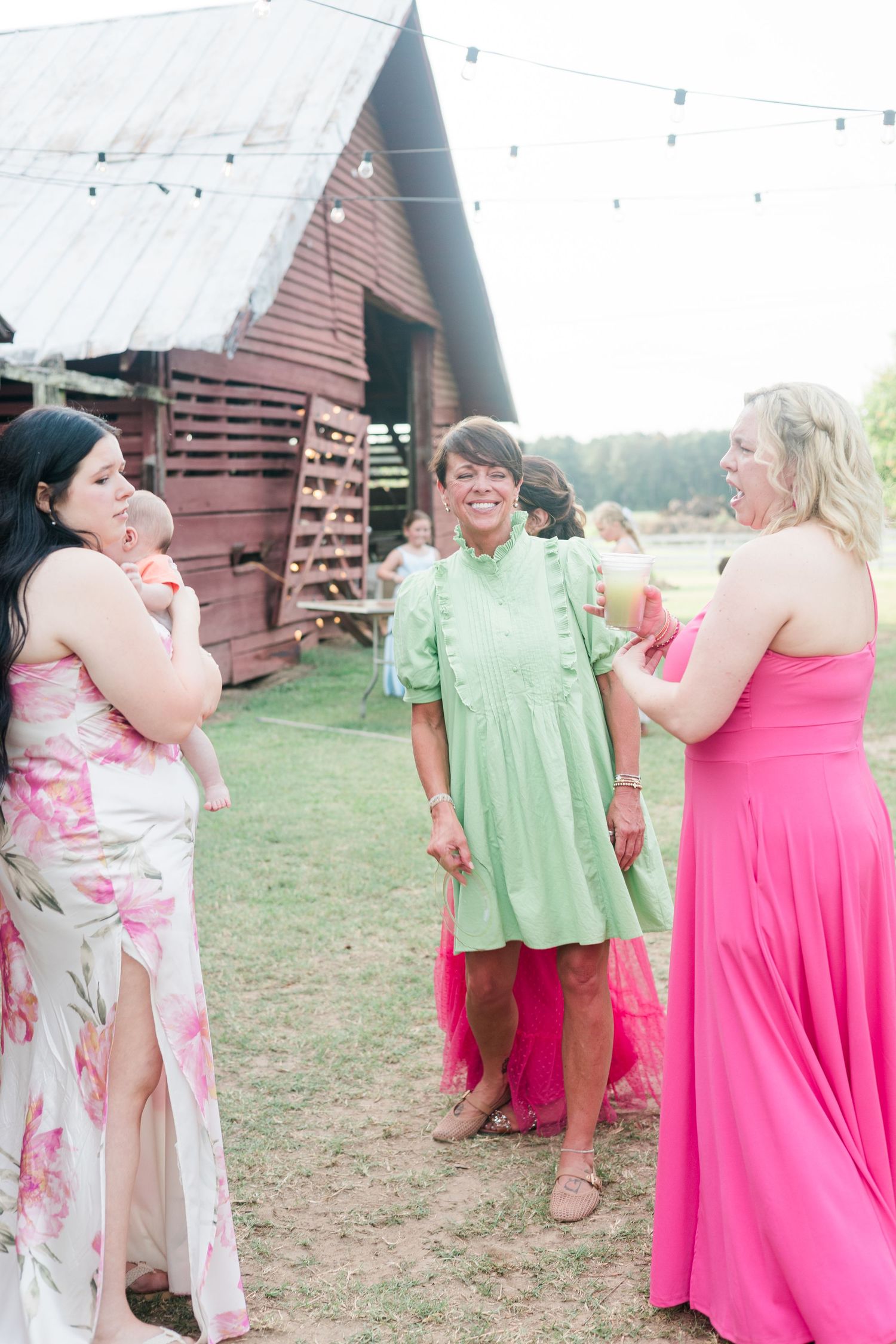 Women in colorful dresses laughing and socializing outdoors near a rustic red barn at a summer wedding reception.