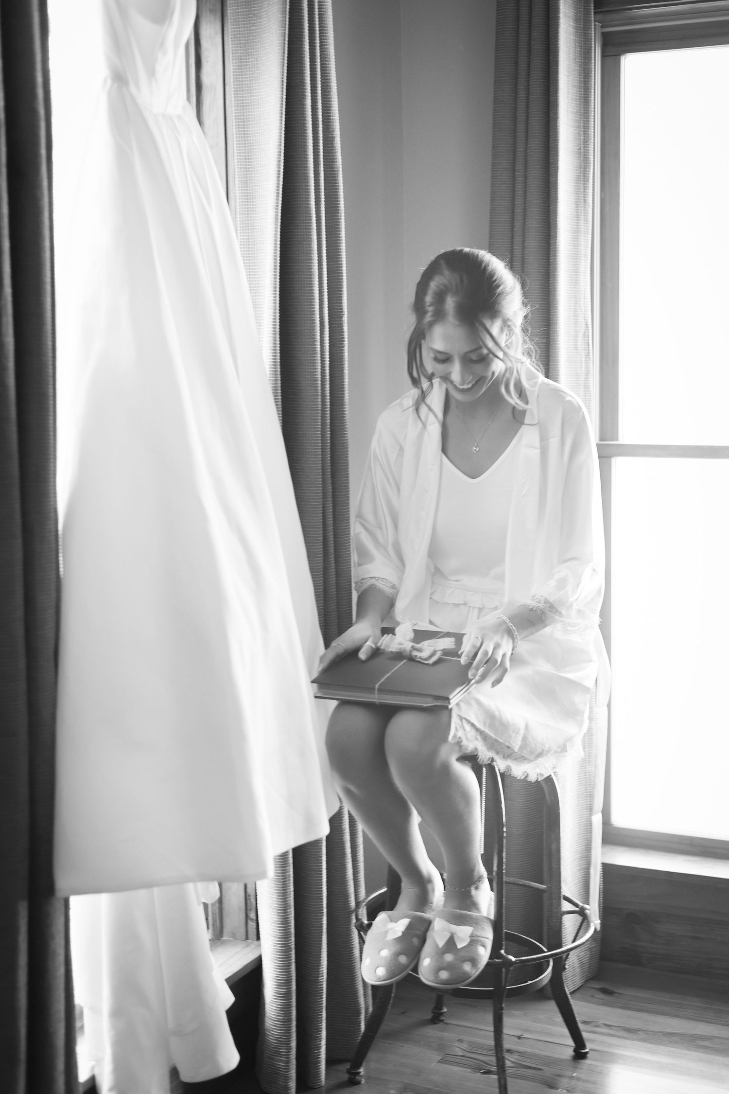 Bride in white robe smiling while opening a gift box, seated by window with wedding dress hanging nearby.