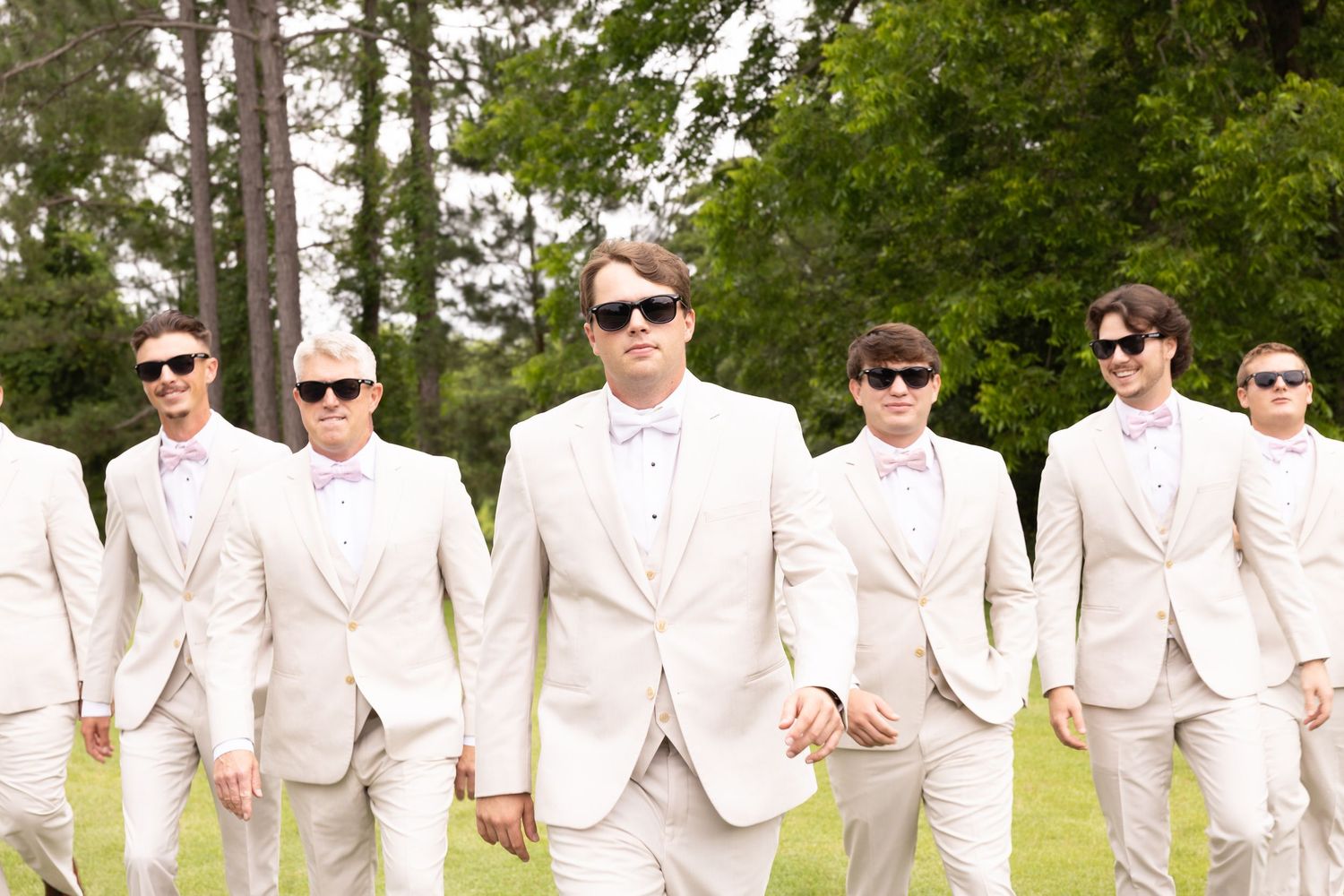 Groomsmen in matching cream suits and sunglasses walking outdoors at a wedding.