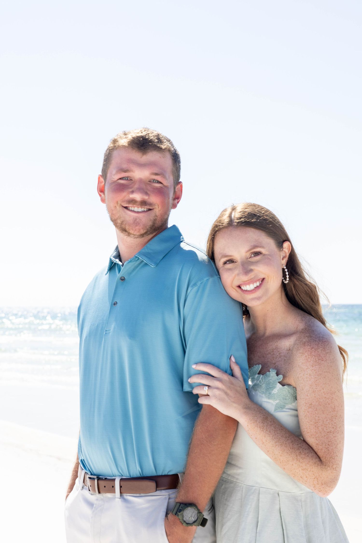Smiling couple posing on sunny beach, man in blue polo shirt with woman in strapless mint dress embracing him.