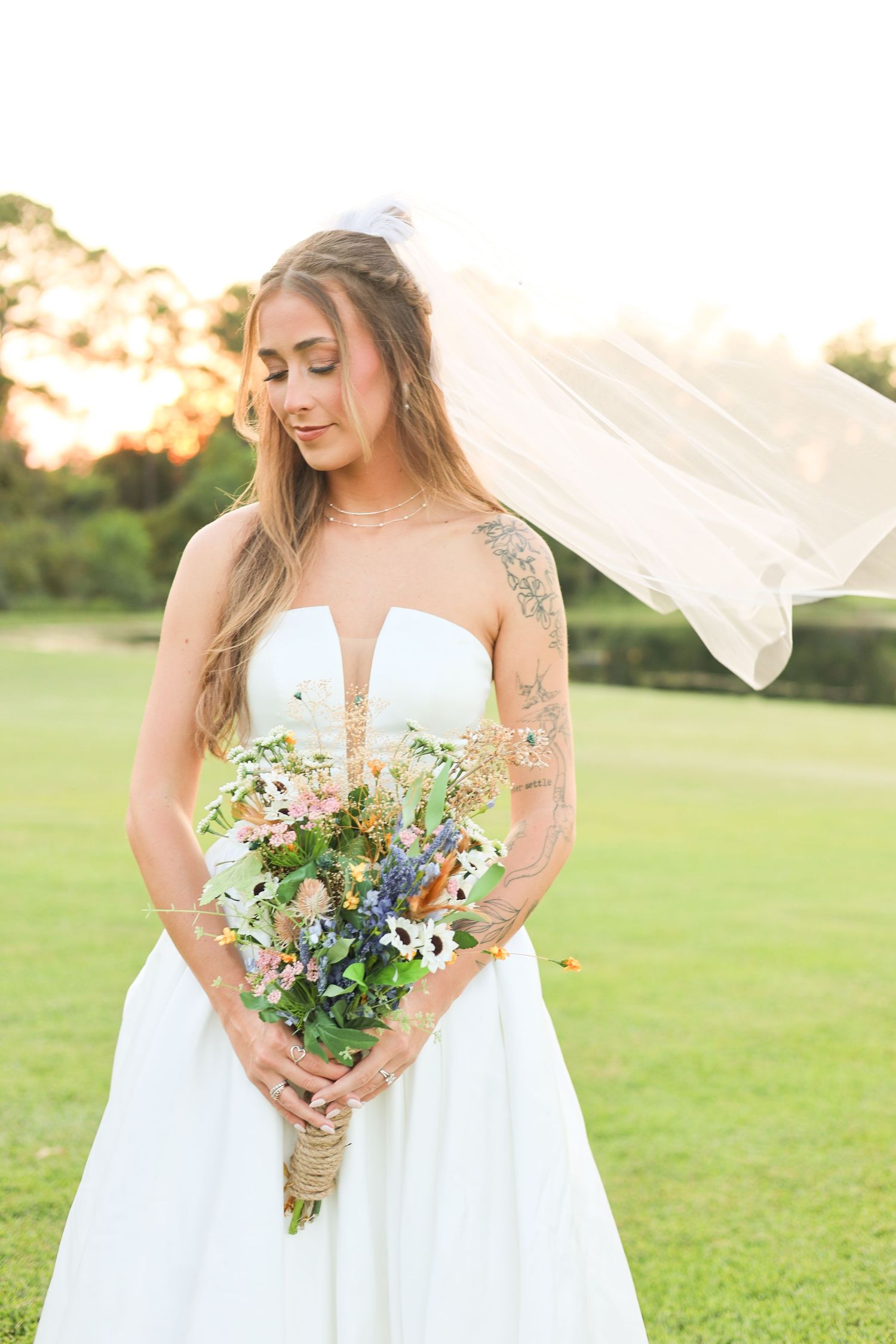 Bride in strapless white gown holding wildflower bouquet, veil flowing in breeze, tattoos visible, golden hour outdoor setting.