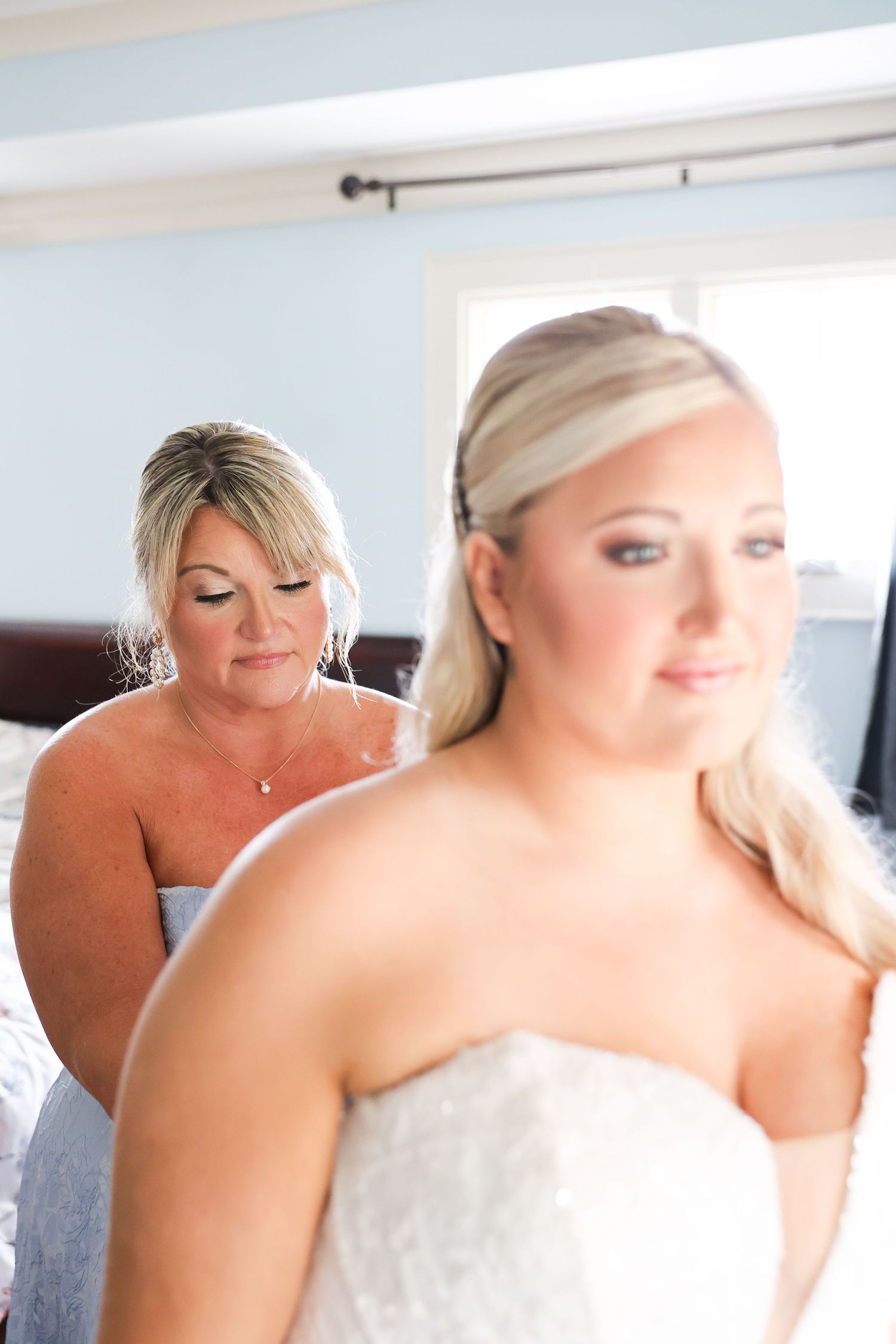 A bride in a strapless white gown stands in focus while a woman helps with her dress in a softly lit bedroom.