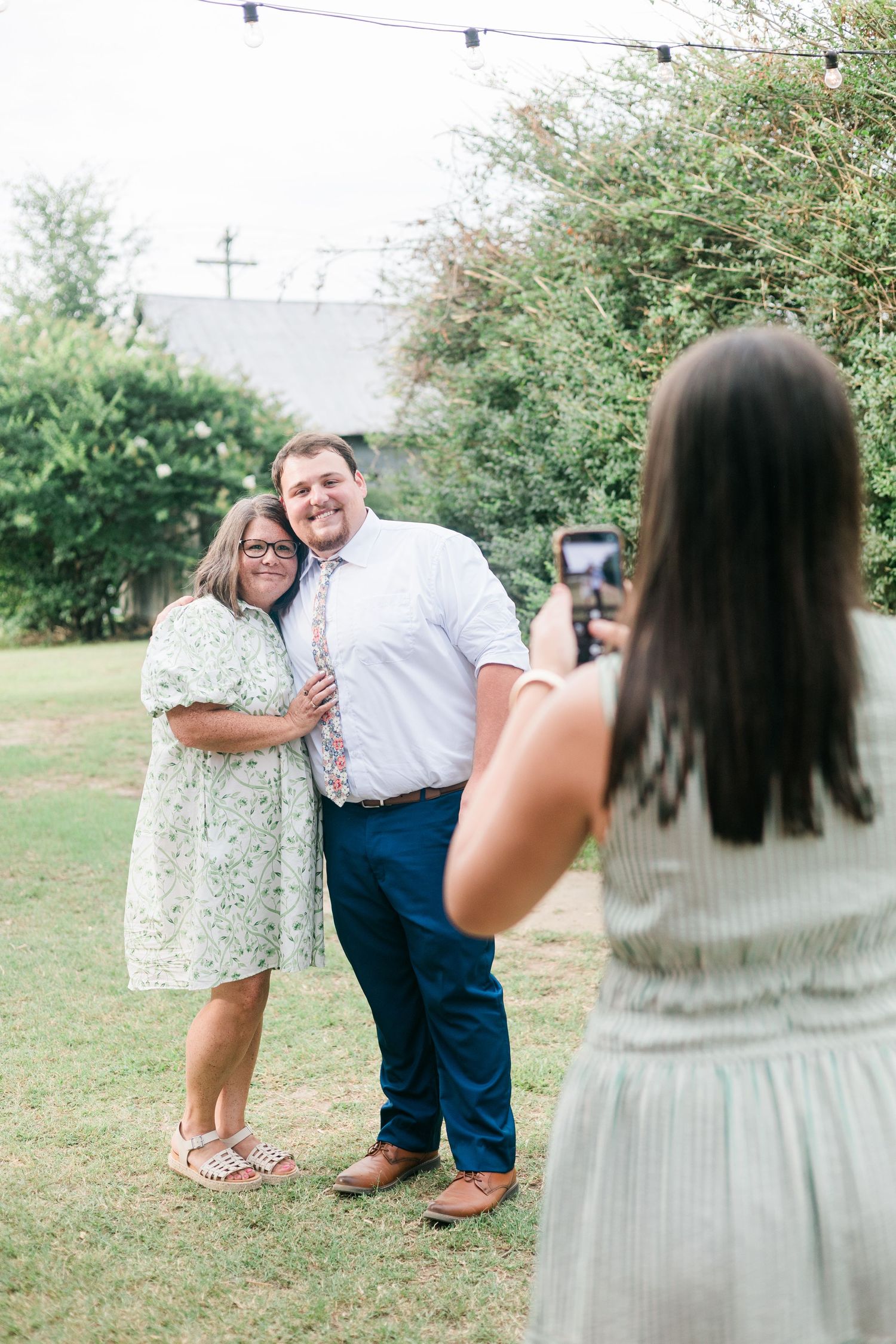 Wedding guest photographs a smiling couple posing together on a green lawn at an outdoor reception.