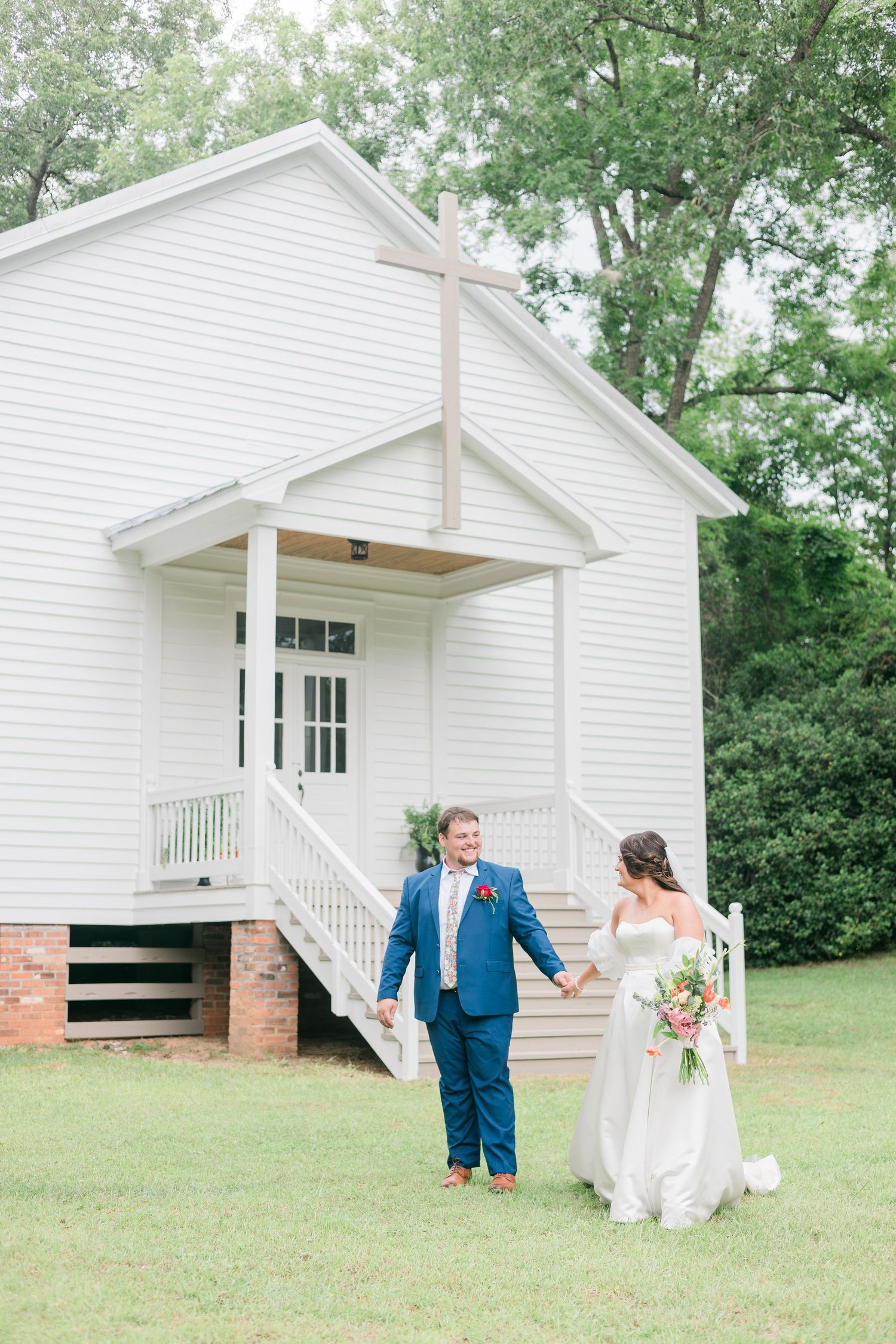 Bride and groom holding hands outside a white chapel with a cross, surrounded by lush green trees on their wedding day.