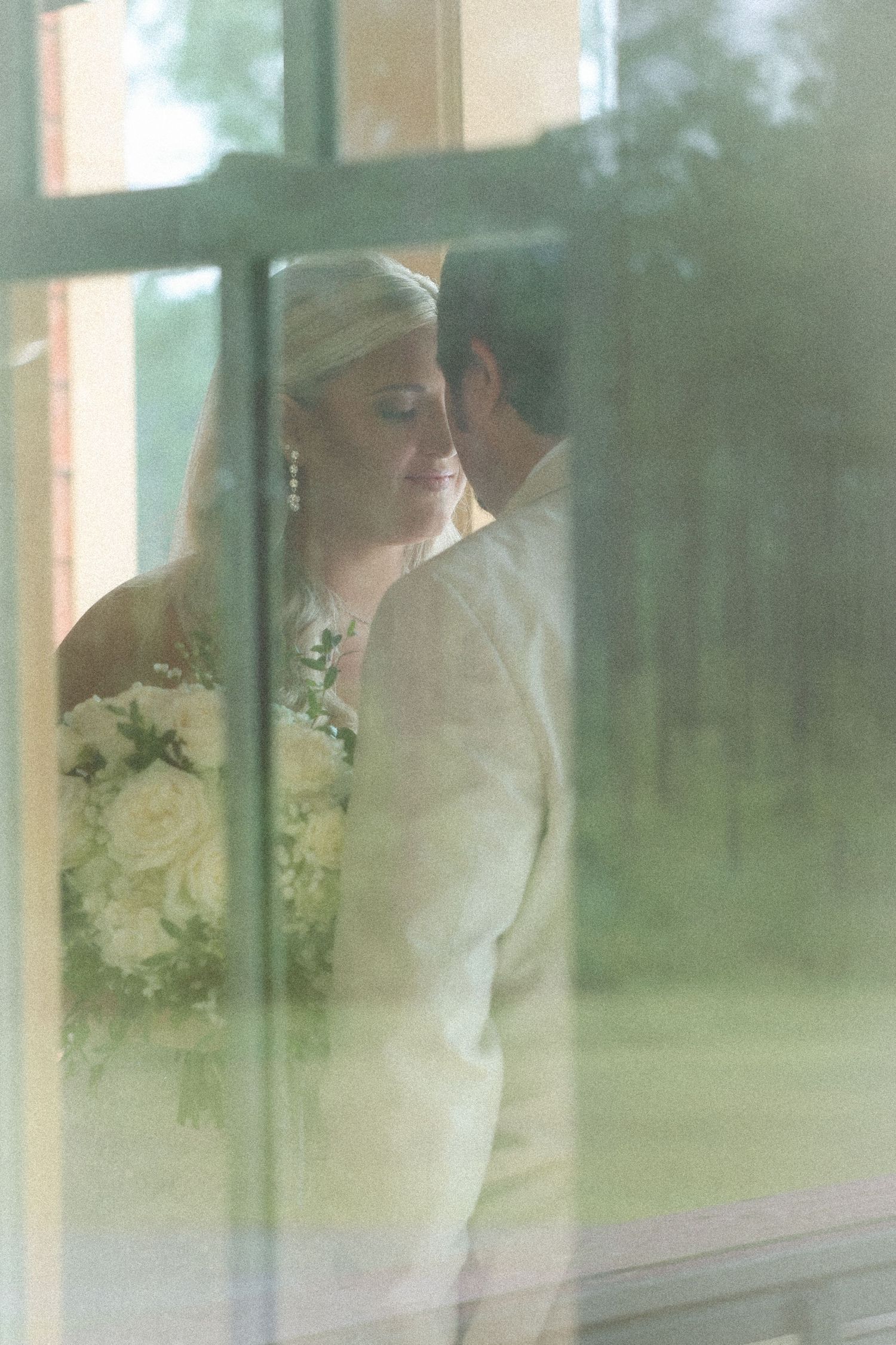 Bride and groom sharing a romantic kiss viewed through a window, bride holding white rose bouquet in wedding attire.