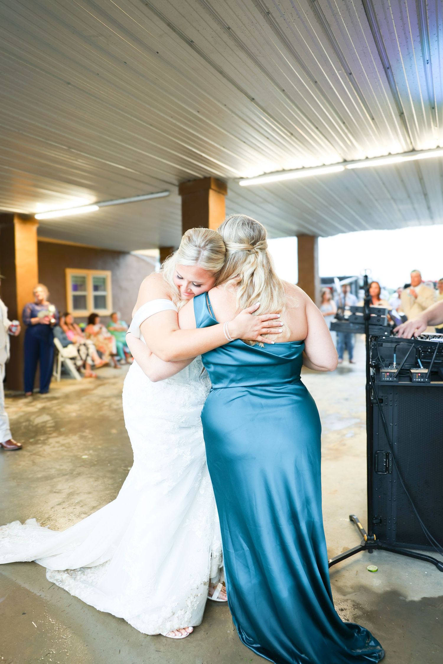 Bride in white lace gown shares emotional hug with bridesmaid in teal satin dress at wedding reception.