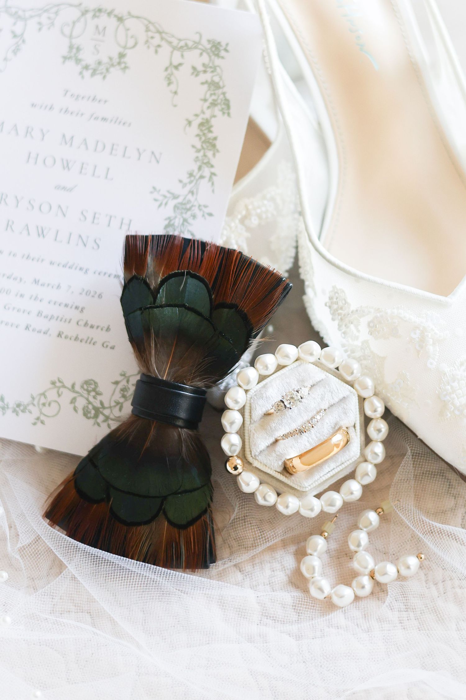 Elegant wedding accessories featuring peacock feather hair clips, pearl bracelets and delicate invitation cards on white lace.